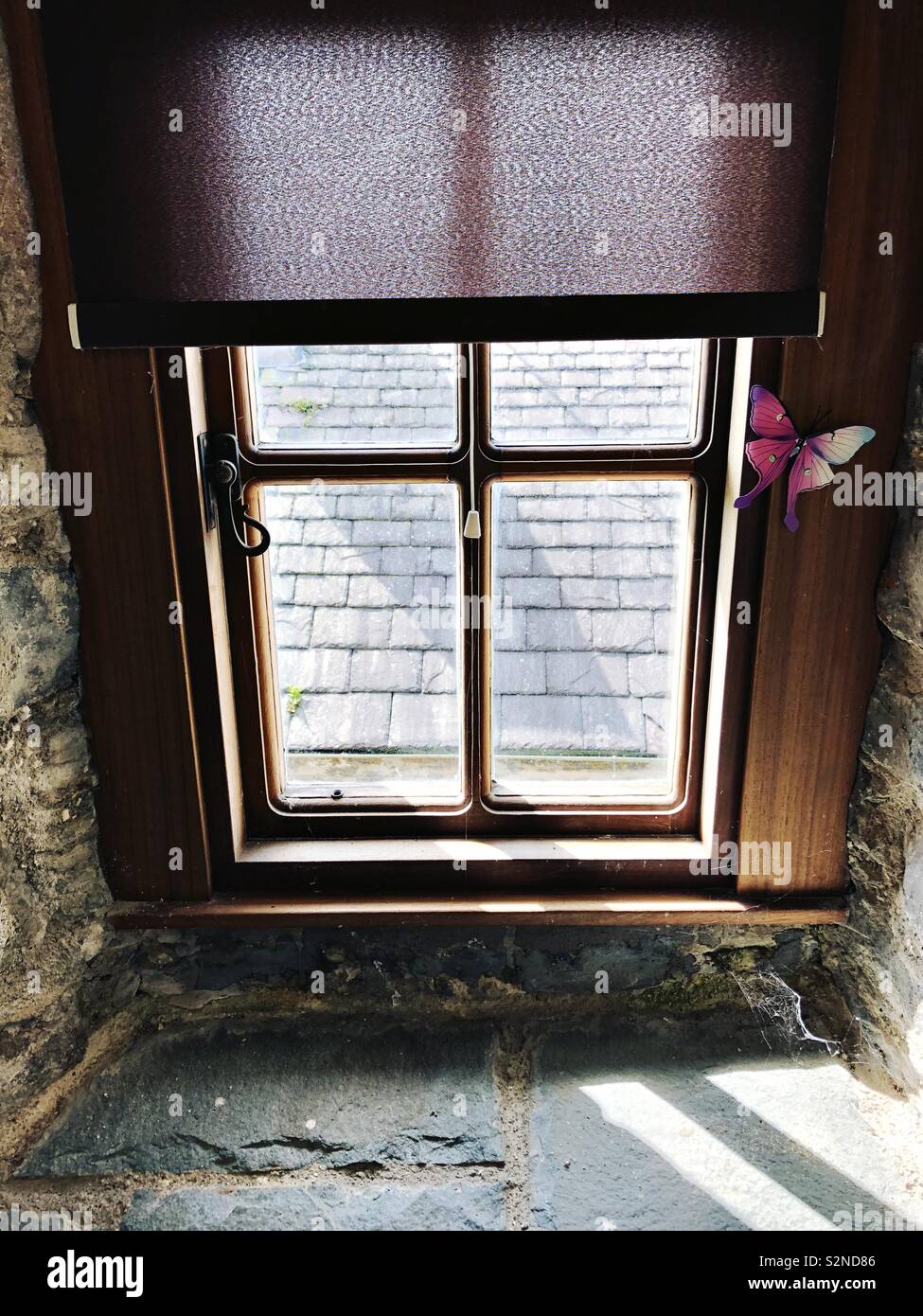 Window in an old building with a dark roller blind and a butterfly ornament.  Old stonework can be seen inside and roof tiles outside. - Smartphone Captured Stock Image