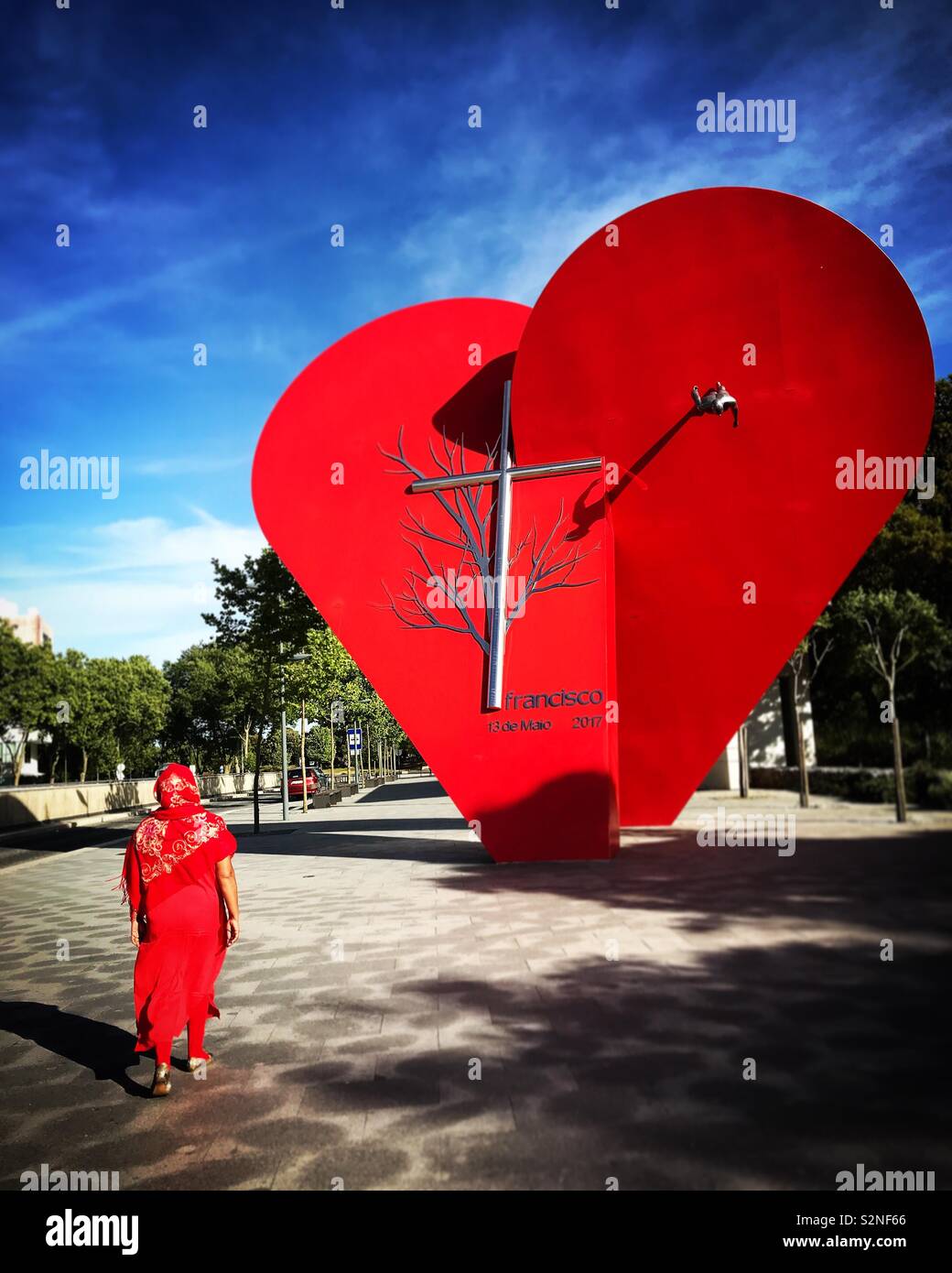A tourist dressed in red visits a monument with of a red heart and a cross in Fatima, Portugal - Smartphone Captured Stock Image