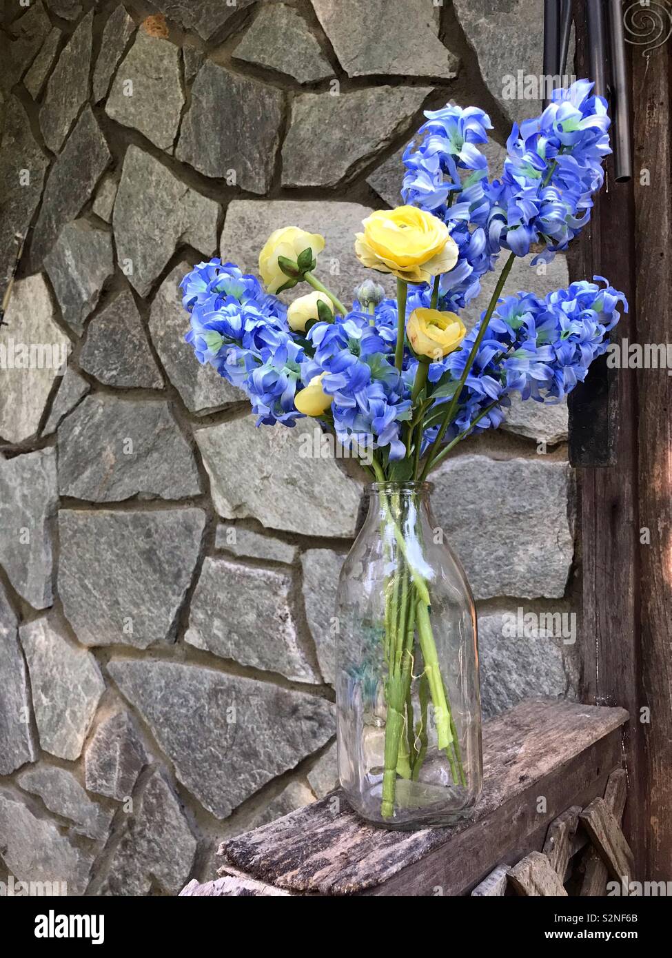 Artificial hyacinth and ranunculus in a clear milk jug sitting on worn wooden railing with stonework in background - Smartphone Captured Stock Image