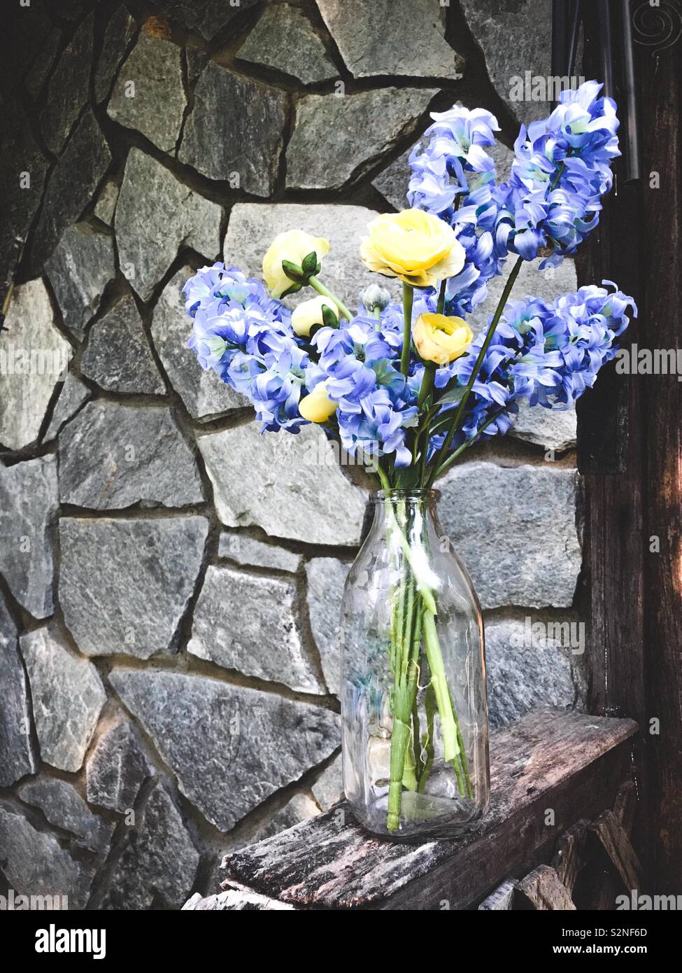 Silk hyacinth and ranunculus in a clear milk jug sitting on worn wooden railing with stonework in background - Smartphone Captured Stock Image