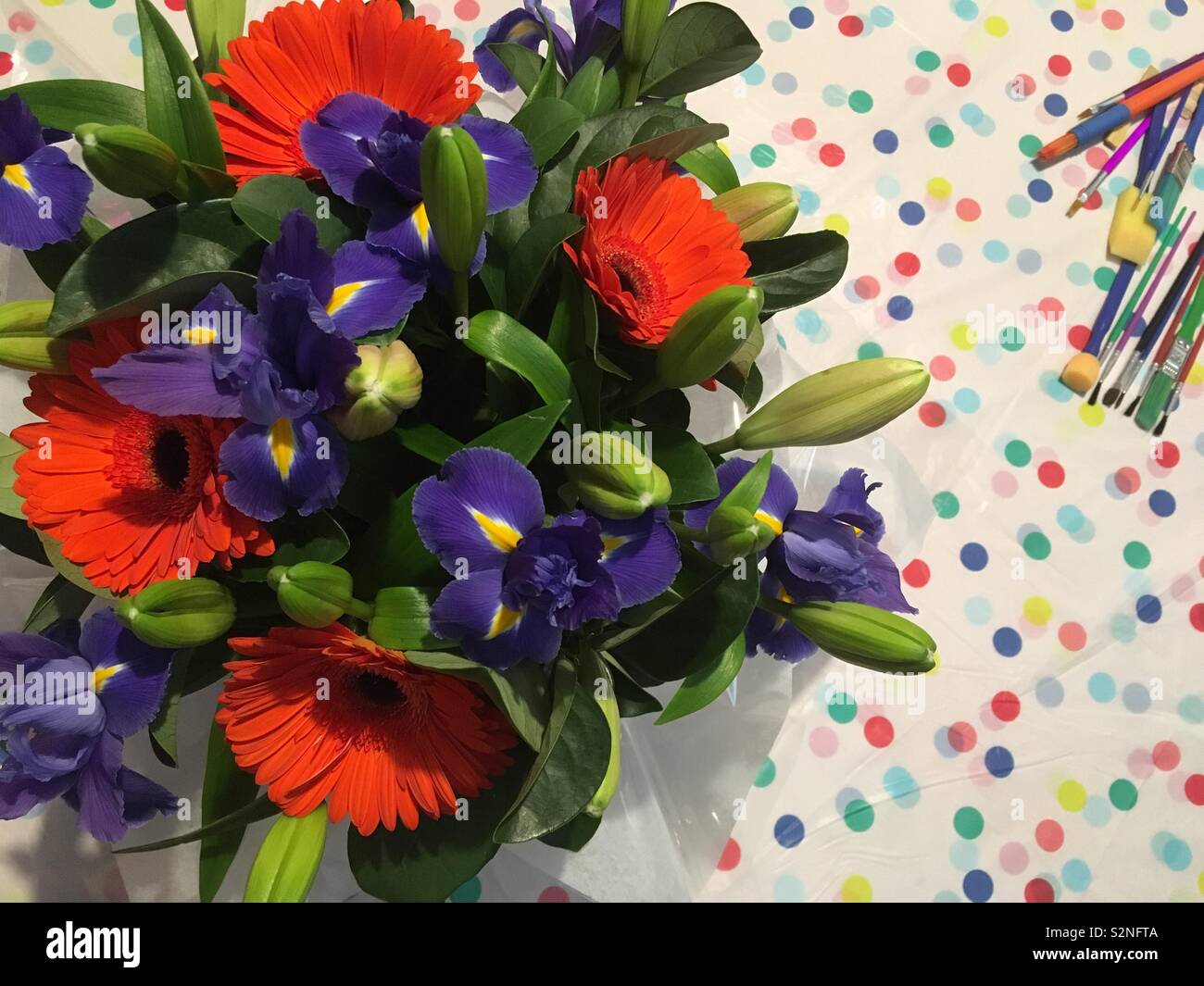 Gerberas, Irises and Lily Buds in a box near artists paint brushes which are all sitting on top of a plastic colourful polka dotted table cover. Stock Photo