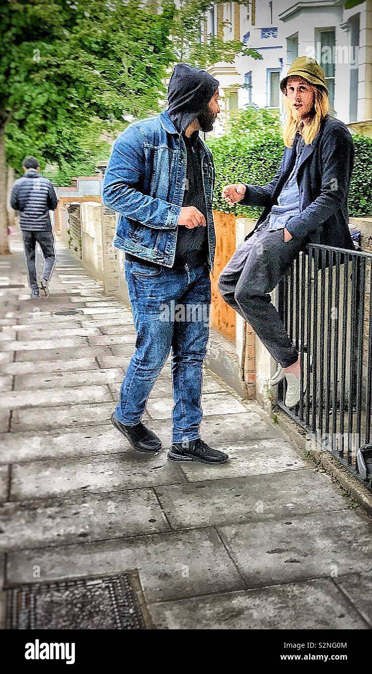 Two young men in casual dress chatting together in the street in Notting Hill district of London, one of whom is sitting on some railings. Stock Photo