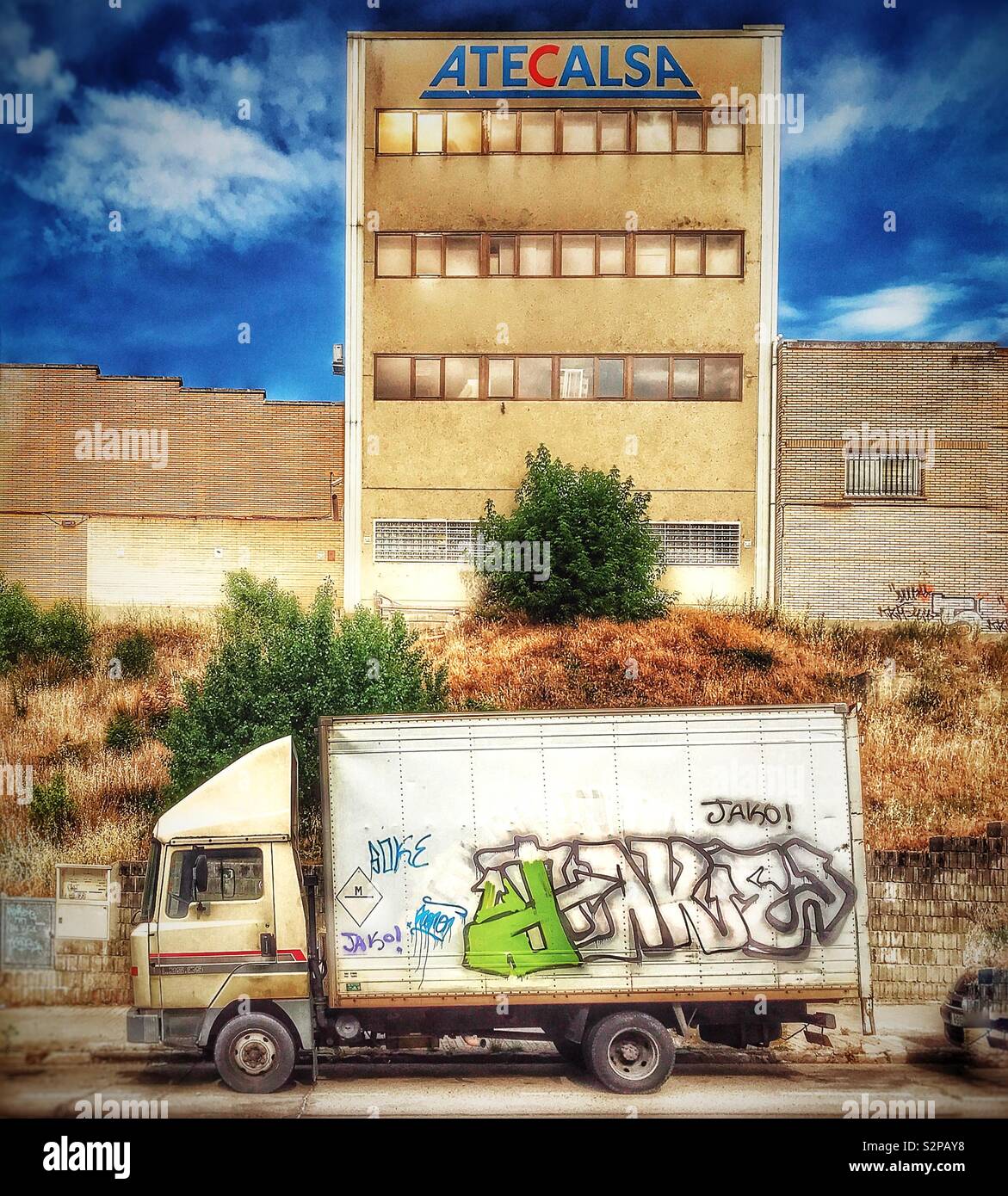 A white lorry and its trailer daubed with graffiti, parked in front of towering buildings on an industrial estate. Stock Photo