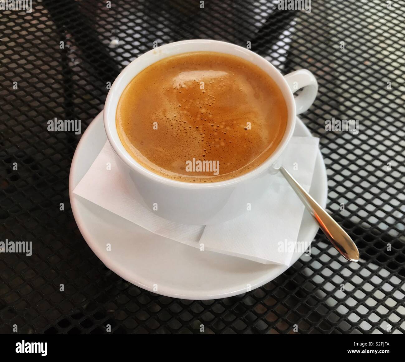 Cup of hot coffee in a while cup and saucer on a black metal mesh table top in an outdoor cafe - Smartphone Captured Stock Image