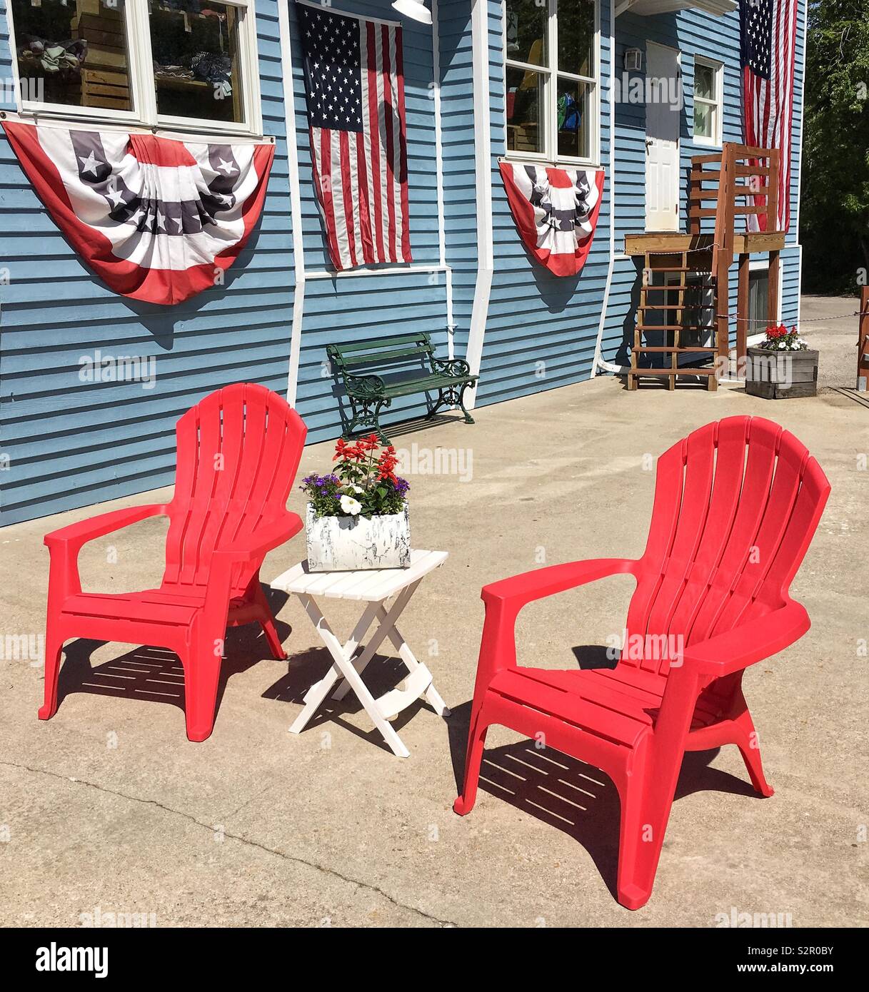 Fourth of July celebration outdoor setup with red Adirondack chairs, American flags and American flag bunting and banners with small side table and red white and blue floral arrangement - Smartphone Captured Stock Image