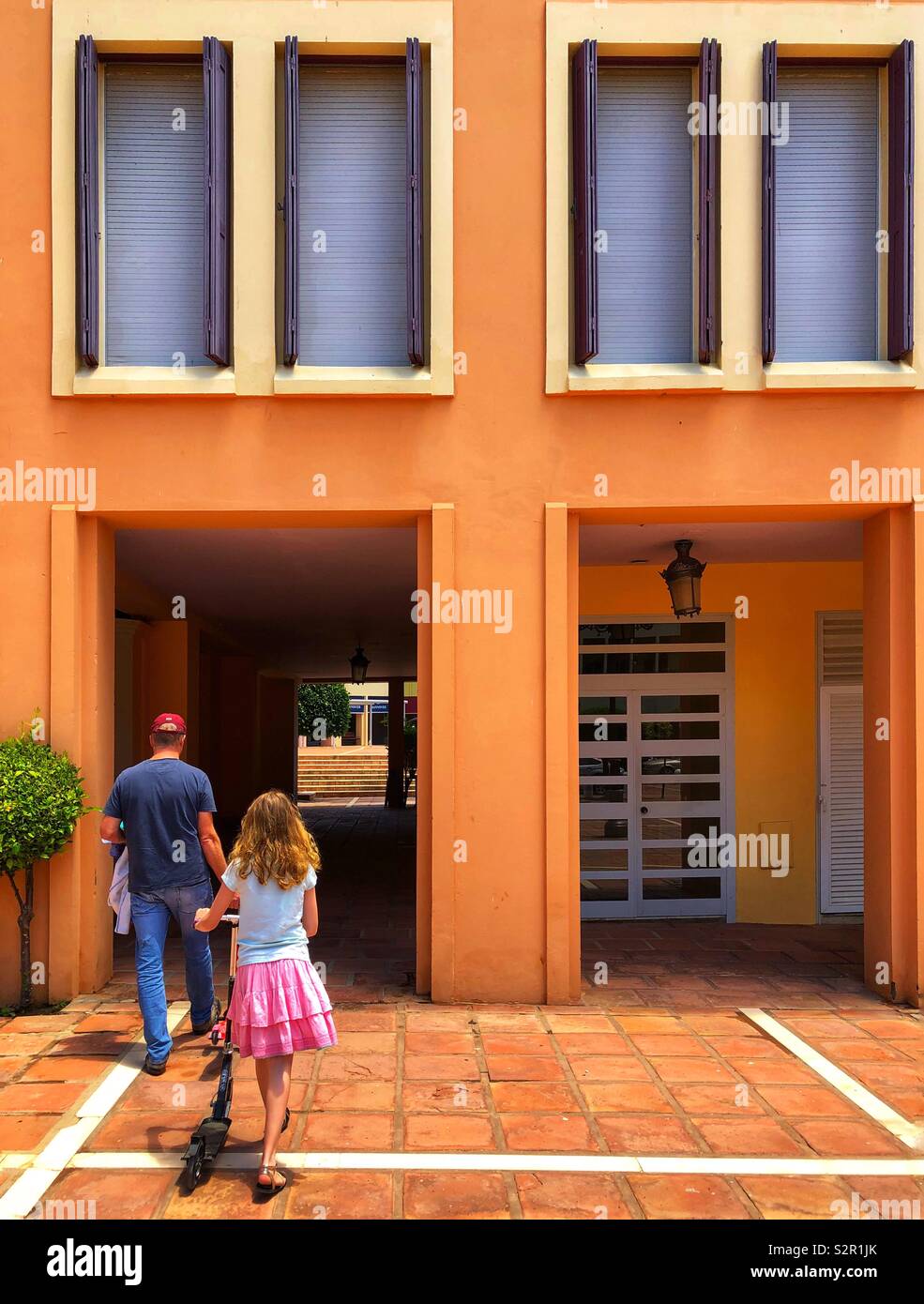 Father and daughter walking into passage under orange building - Smartphone Captured Stock Image