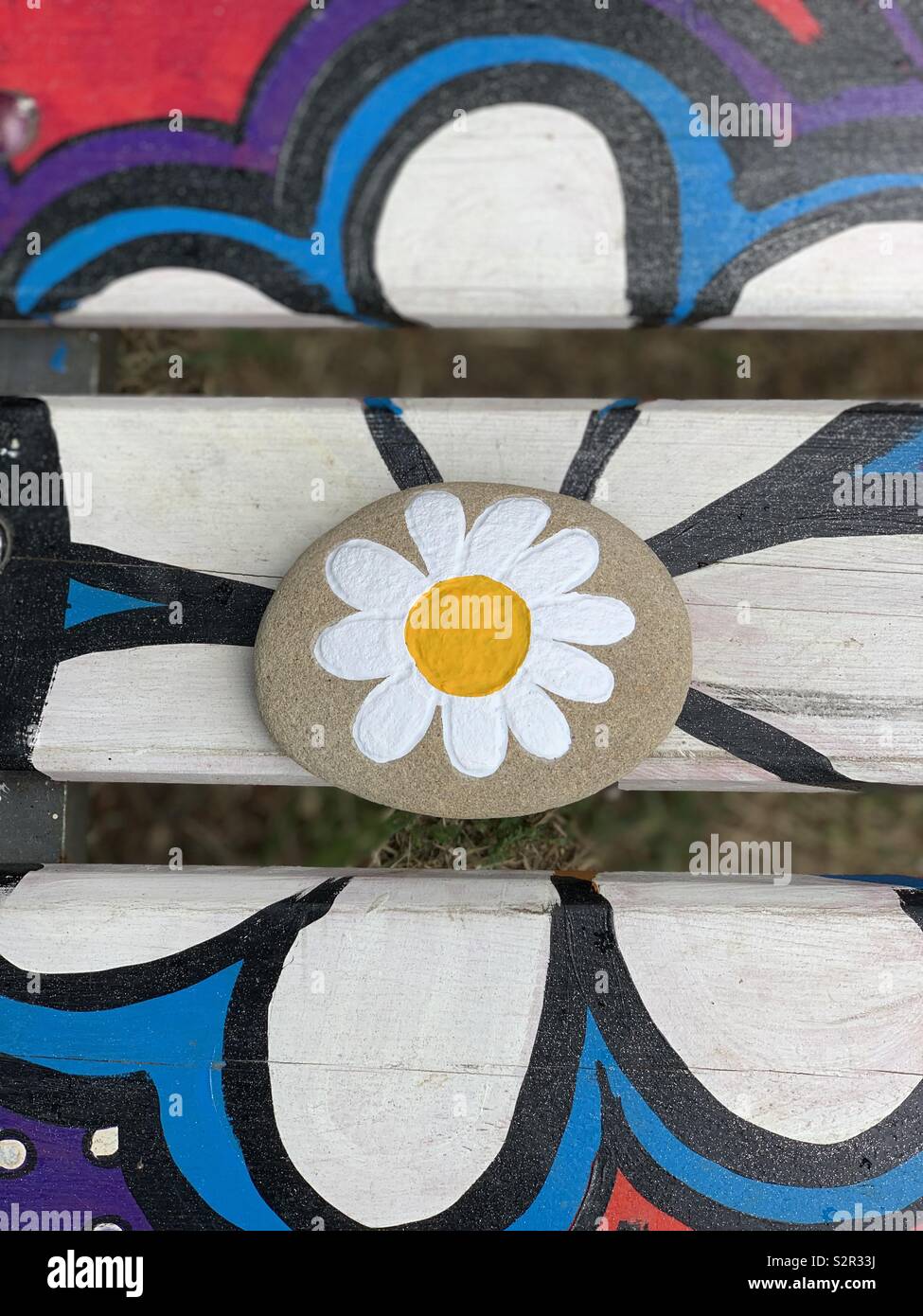 Peace message with a daisy design on a stone over a painted daisy over a public bench Stock Photo
