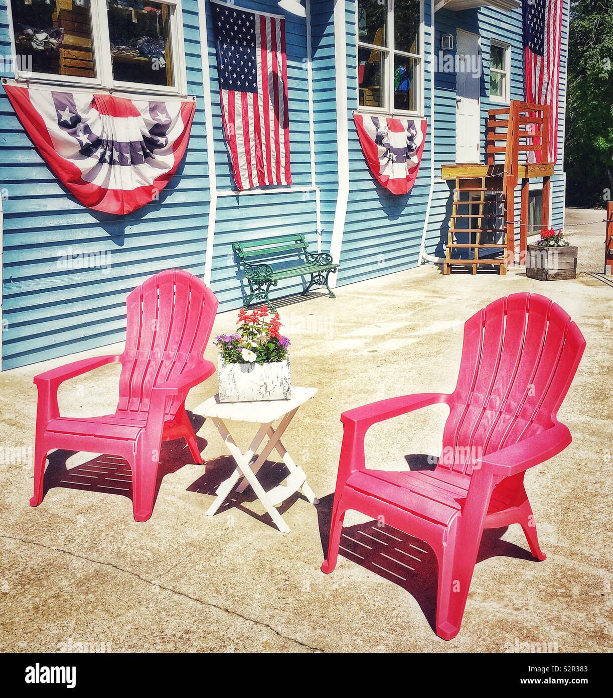Fourth of July celebration outdoor setup with red Adirondack chairs, American flags and American flag bunting and banners with small side table and red white and blue floral arrangement - Smartphone Captured Stock Image