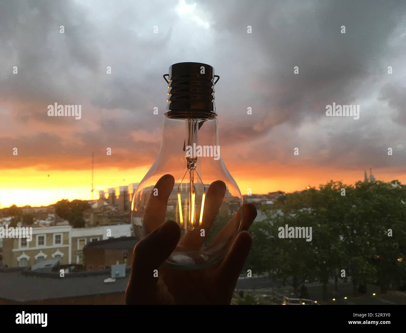 Hand holding up solar bulb against sunset and storm clouds - Smartphone Captured Stock Image