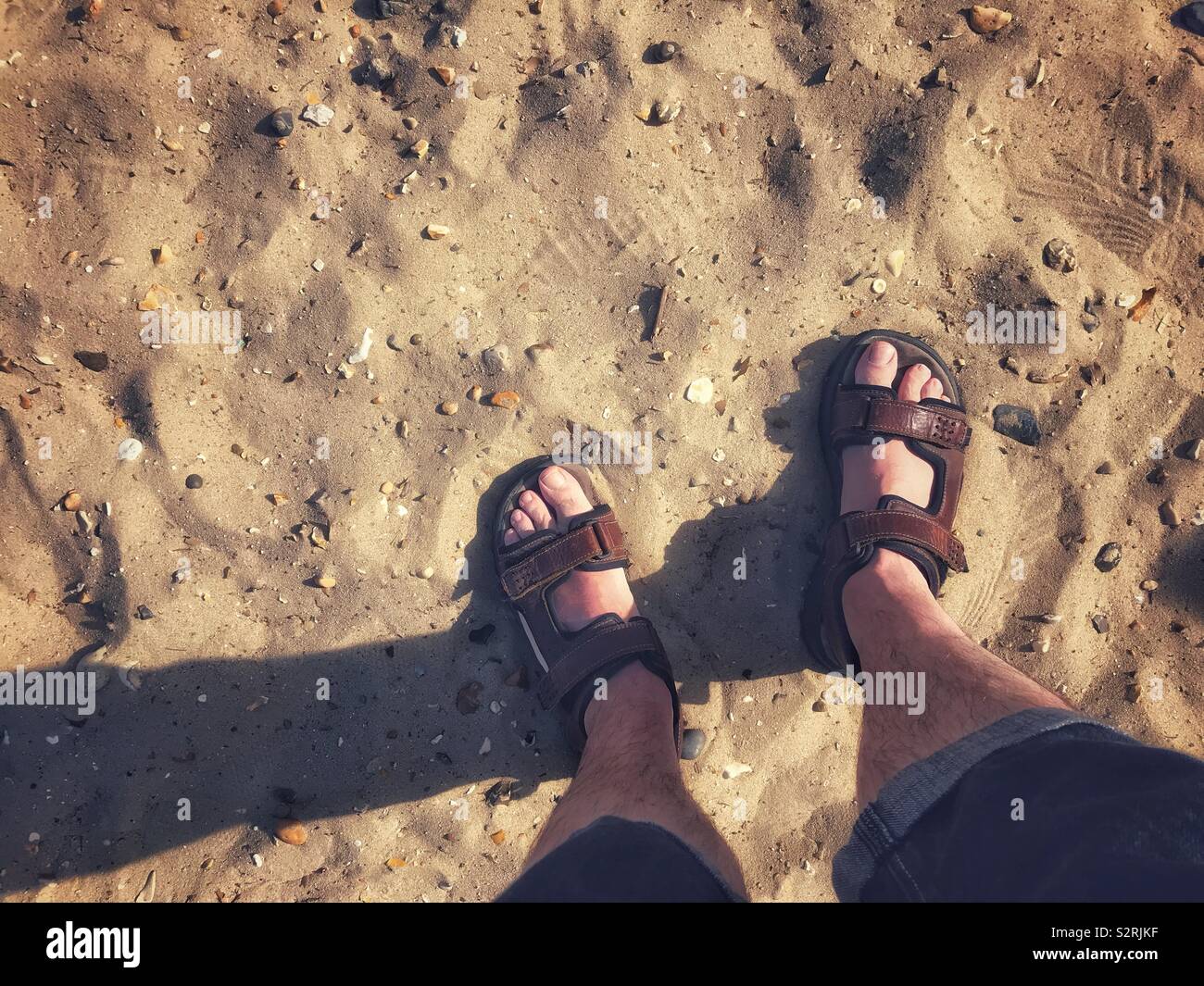 A pair of male feet wearing sandals on a sandy beach. Stock Photo