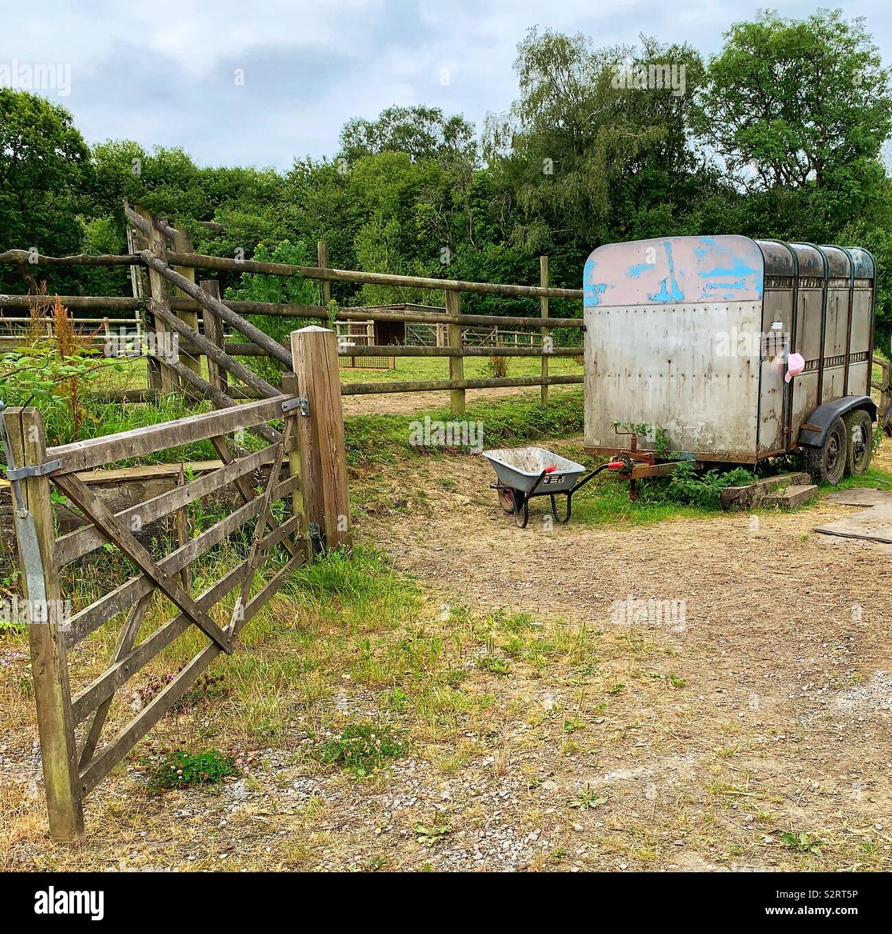 Open wooden gate and fence leading to horsebox with wheelbarrow Stock Photo