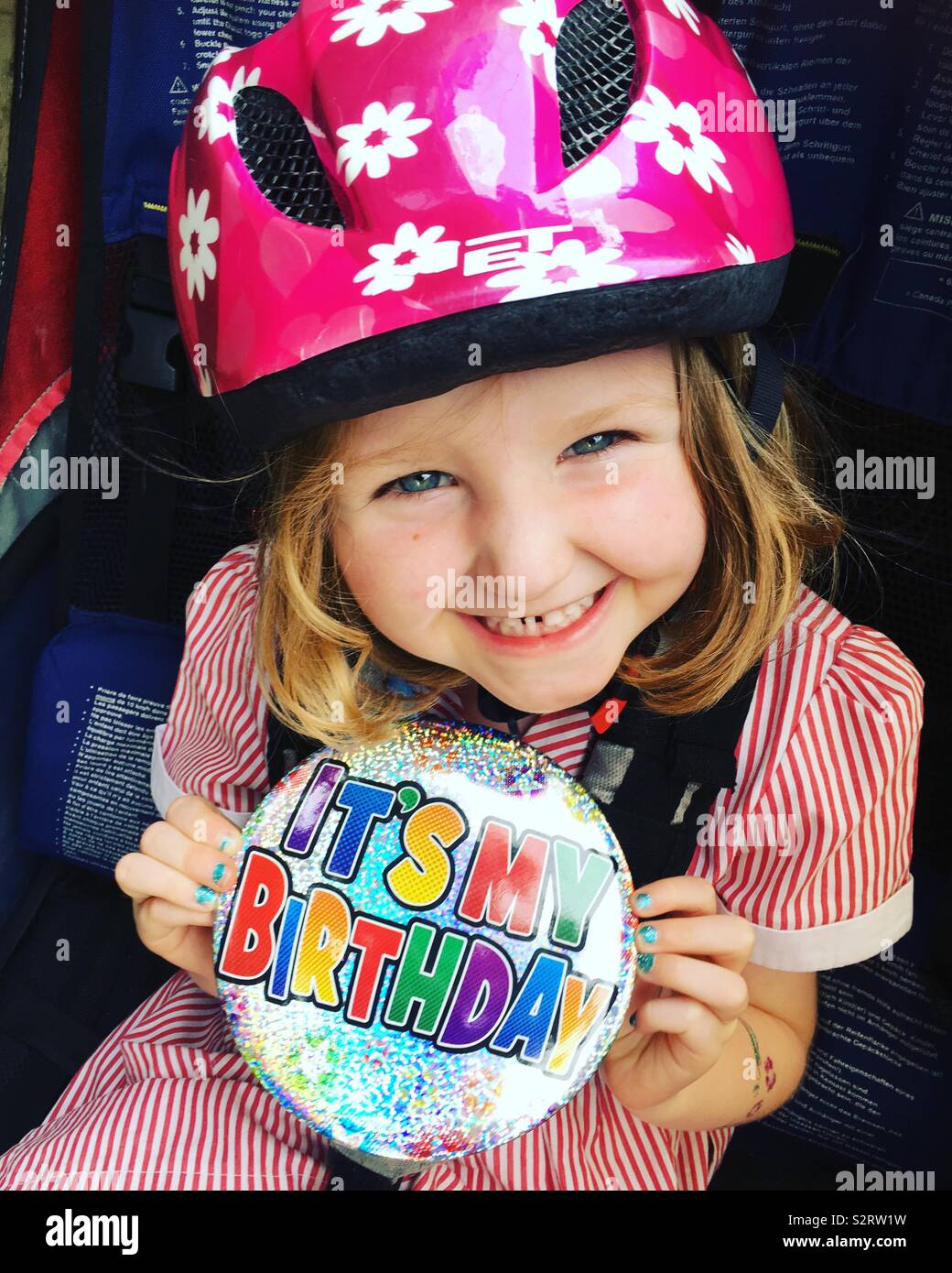 Five-year-old girl with a smile and wearing an oversize / oversized Happy Birthday It’s My Birthday badge / large big badge while sitting in a bicycle bike trailer and wearing a cycle helmet / hat - Smartphone Captured Stock Image