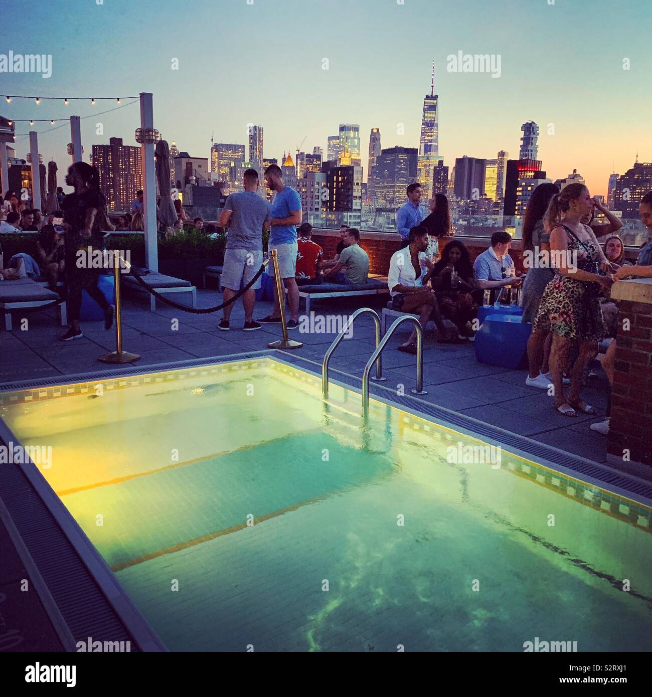 Patrons of the Mr. Purple bar socialize near the pool with the Lower Manhattan skyline in the background at the Hotel Indigo Lower East Side, New York City - Smartphone Captured Stock Image