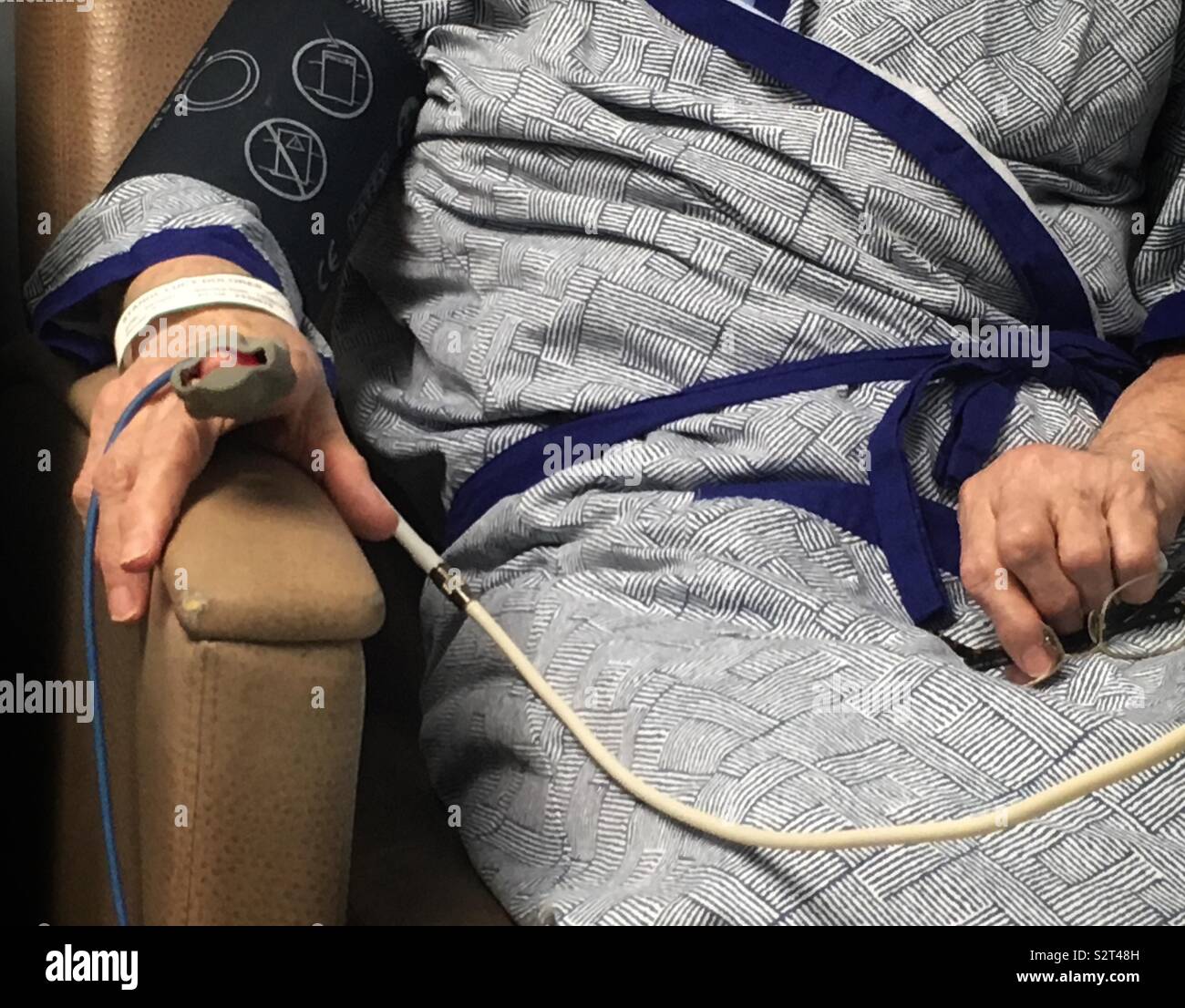 Closeup of a seniors mid-section, and hands, one hand with heart monitor attached - wearing hospital gown while seated in chair Stock Photo