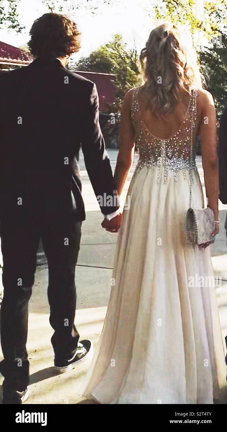 Prom couple with backs showing, walking while holding hands wearing a tuxedo and tennis shoes and beautiful blonde wearing cream dress in the early evening Stock Photo