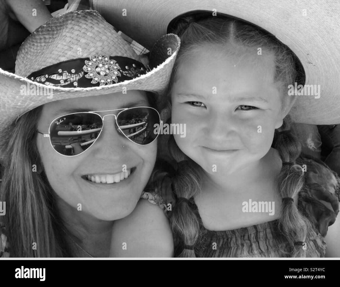 Woman wearing aviator sunglasses and young girl, both wearing cowboy hats while at the rodeo smiling. Black and white photo. Stock Photo