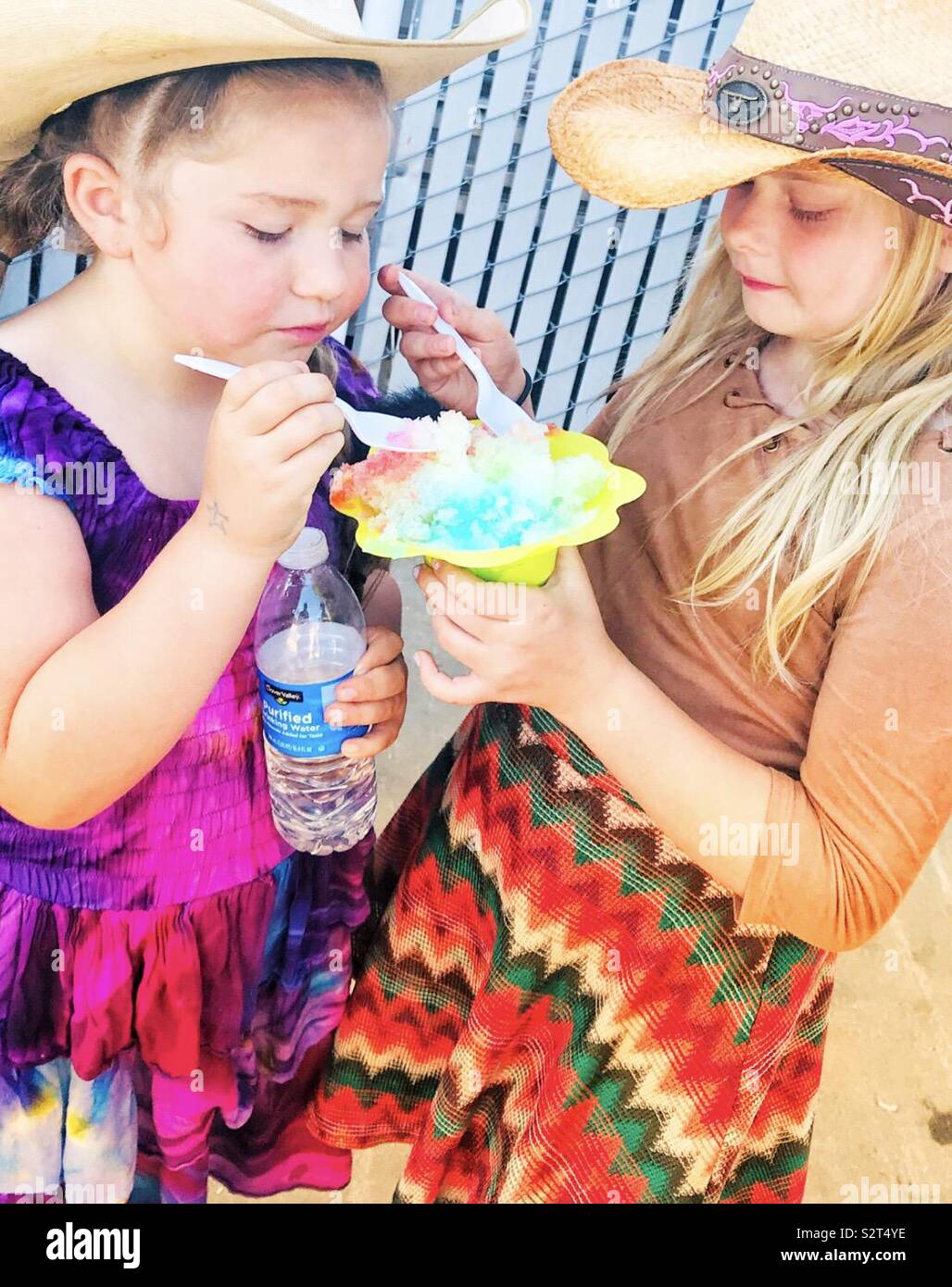 Two young girls wearing cowboy hats at the rodeo, sharing a huge snow cone wearing bright dresses Stock Photo