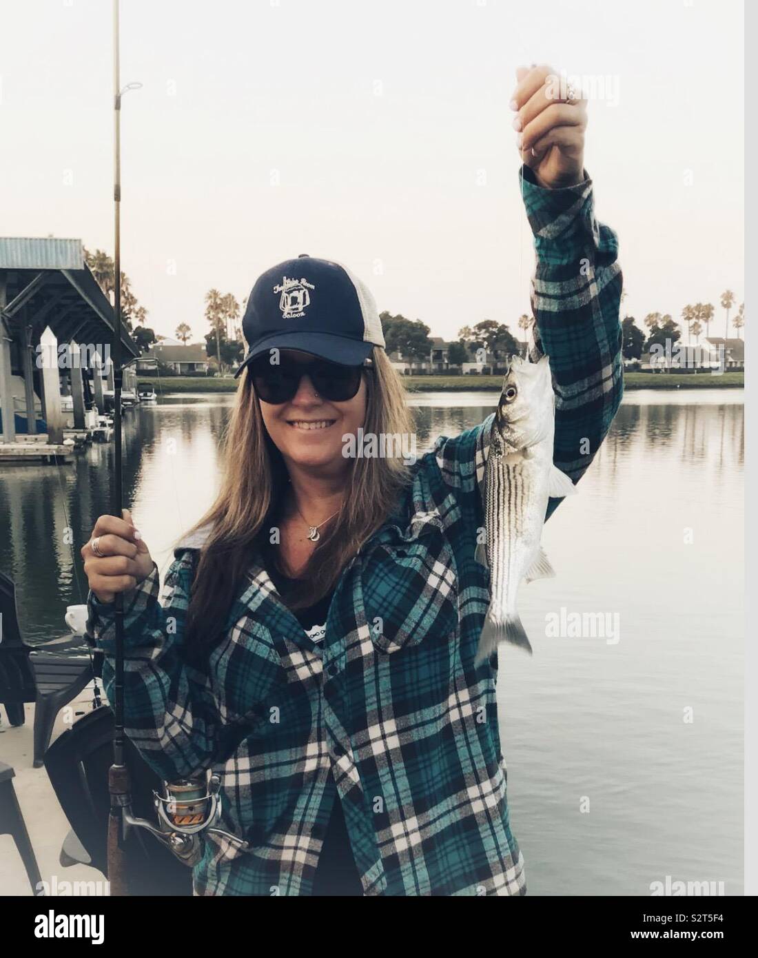 Woman wearing a flannel and trucker hat, just caught a fish, holding the fish in the line, water, trees and Marina in background, daytime Stock Photo