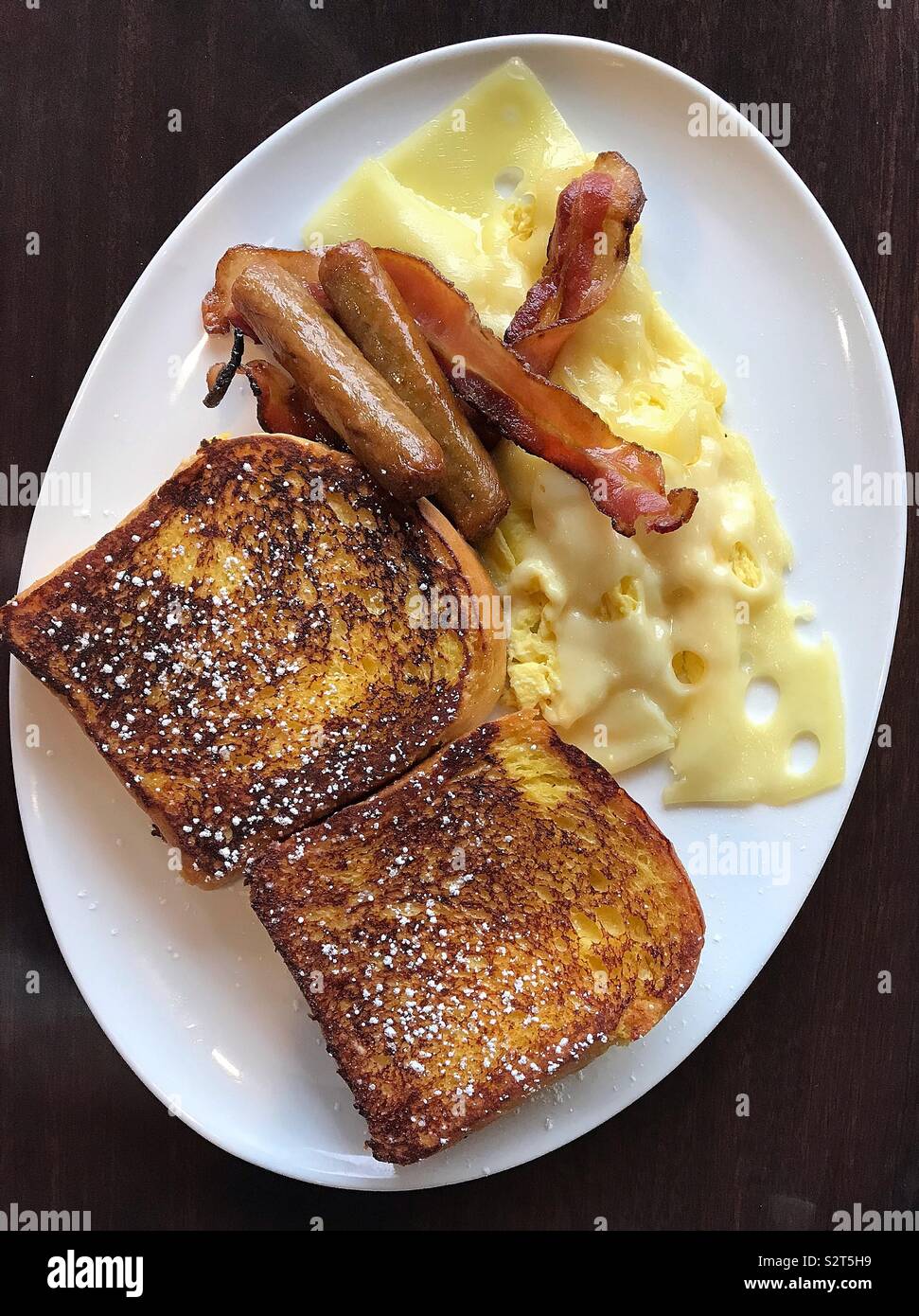 A mouth-watering meal of fluffy French toast, crispy bacon, sausages, and a Swiss cheese omelette sits on a large white oval plate. The bright meal sits on a dark brown table. Stock Photo
