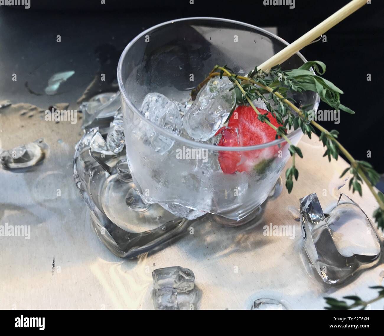 Cocktail glass garnished with strawberry, thyme, and lemongrass sits broken on a stainless steel surface, surrounded by ice, alcohol, and broken glass. Stock Photo