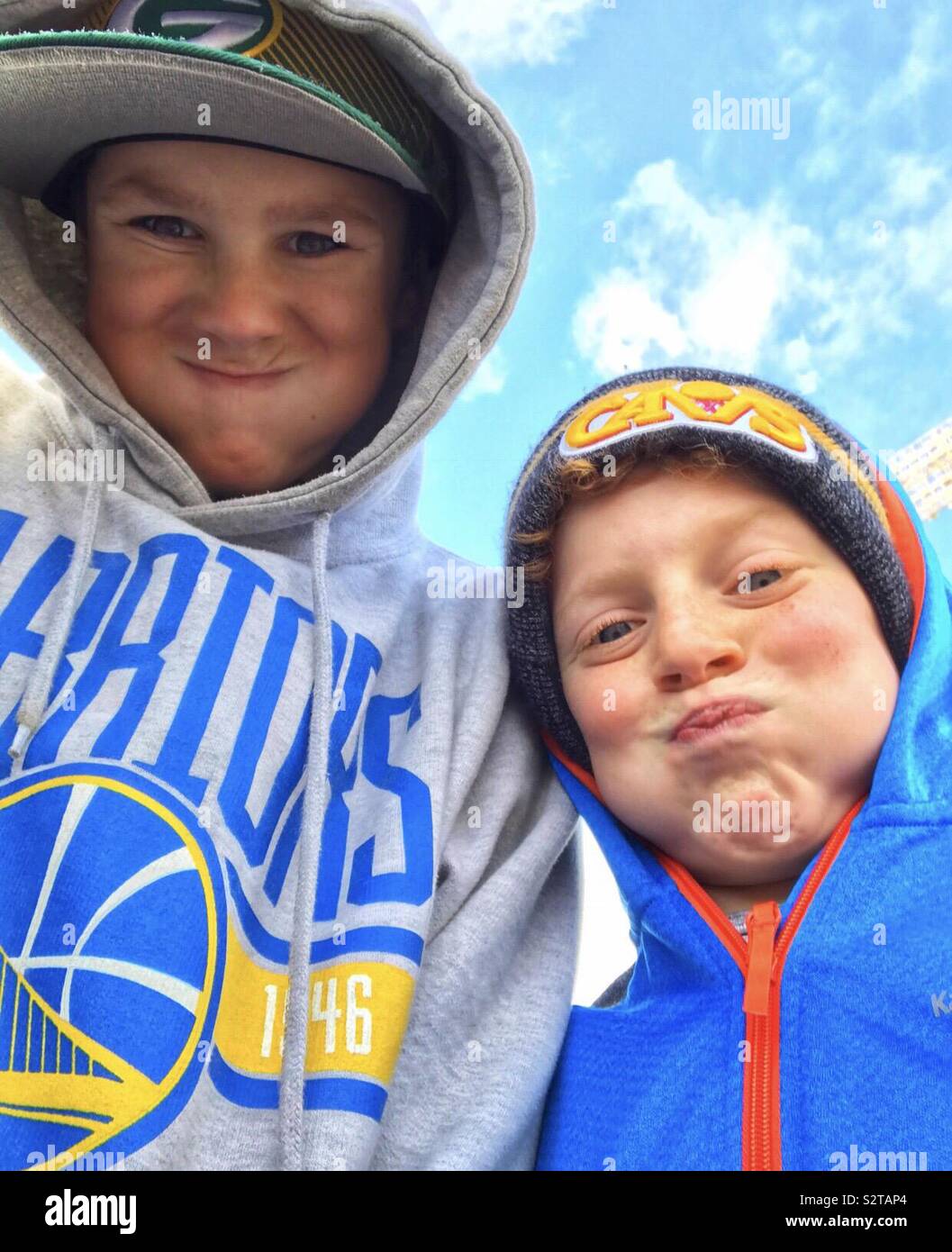Two preteen boys wearing sweatshirts, hats and hoodies on their heads,while making silly faces and puffy cheeks. Bile sky with clouds Stock Photo