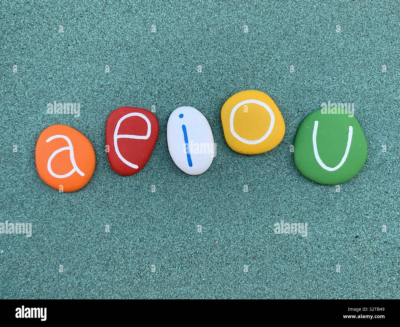 A,e,i,o,u, vocal letters of the alphabet composed with colored stones over green sand Stock Photo