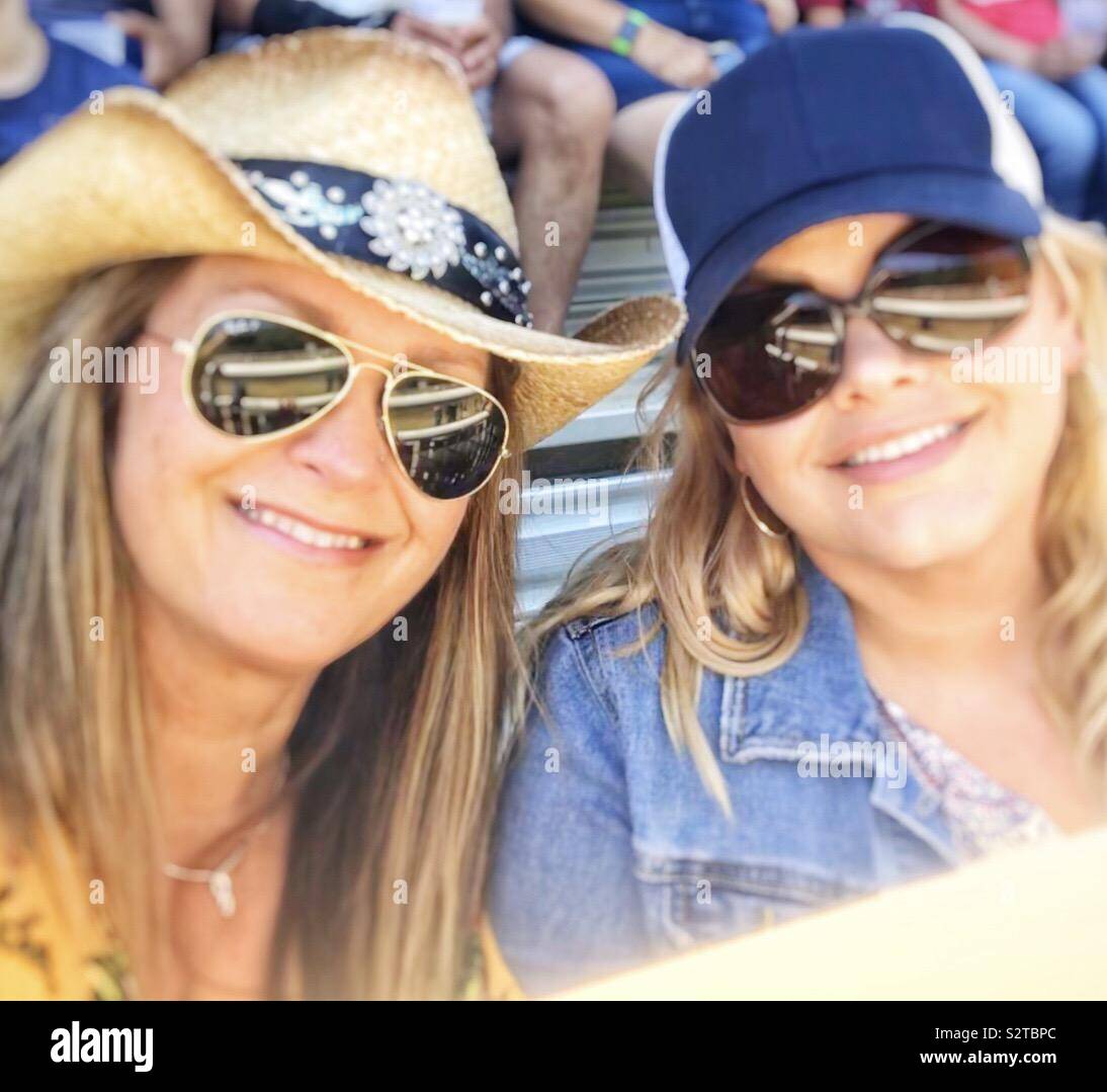 Two women at the rodeo in the stands wearing a cowboy hat, trucker hat and both in sunglasses Stock Photo