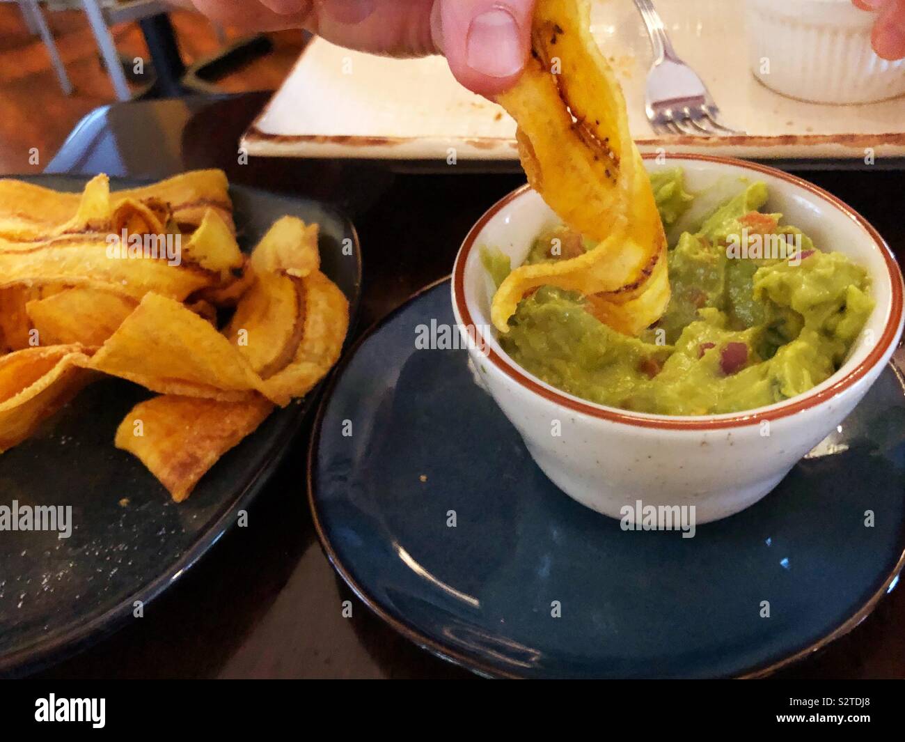 Men’s hand dipping plantain chip into a small bowl of guacamole with a plate of extra chips beside Stock Photo