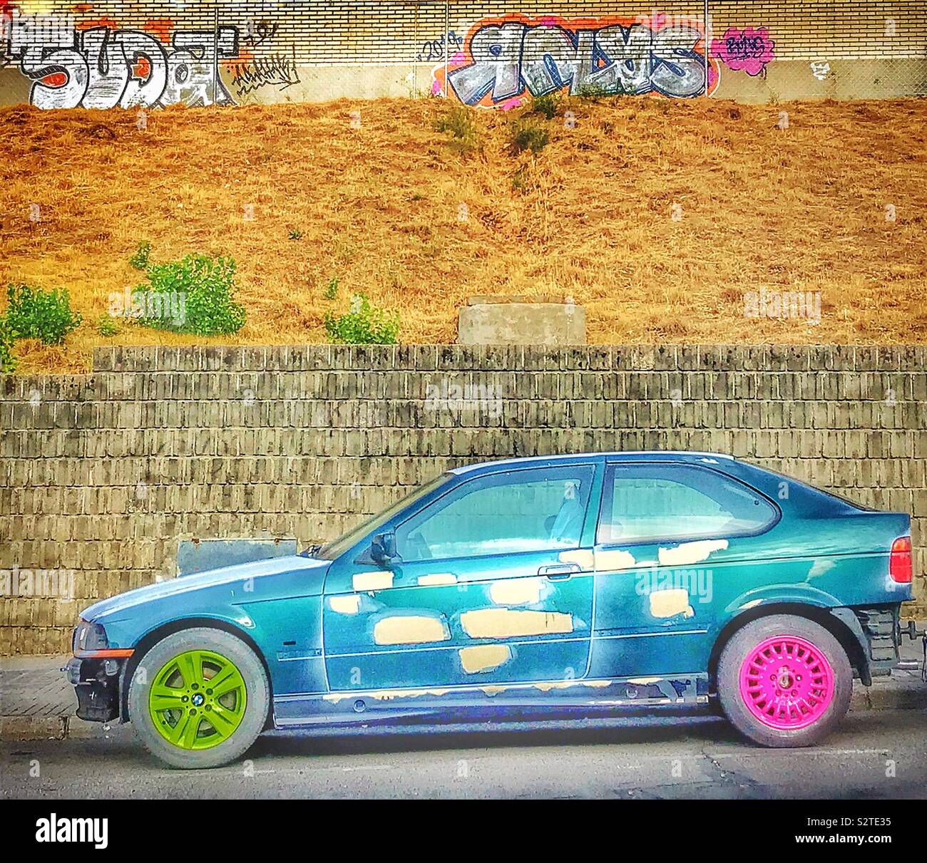 A car has wheels painted in different colours, line green and pink. The bodywork is daubed with splashes of grey paint. It is parked in front of an old building daubed with graffiti. Stock Photo