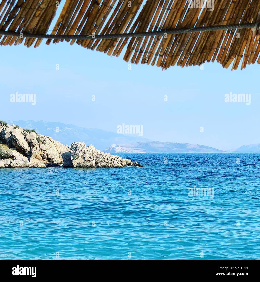 View of sea and cliffs on Dalmatian islands from under a straw beach umbrella. Oprna Bay, Stara Baška, Croatia. - Smartphone Captured Stock Image