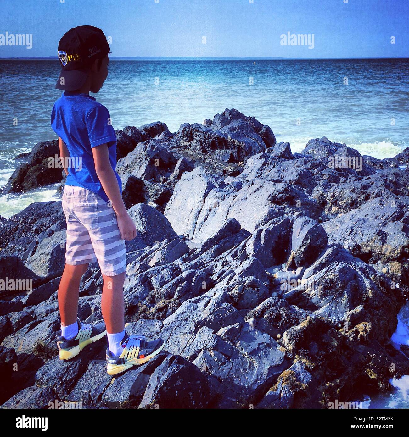 1 young boy looking at the sea or ocean. He stands on rocks Stock Photo