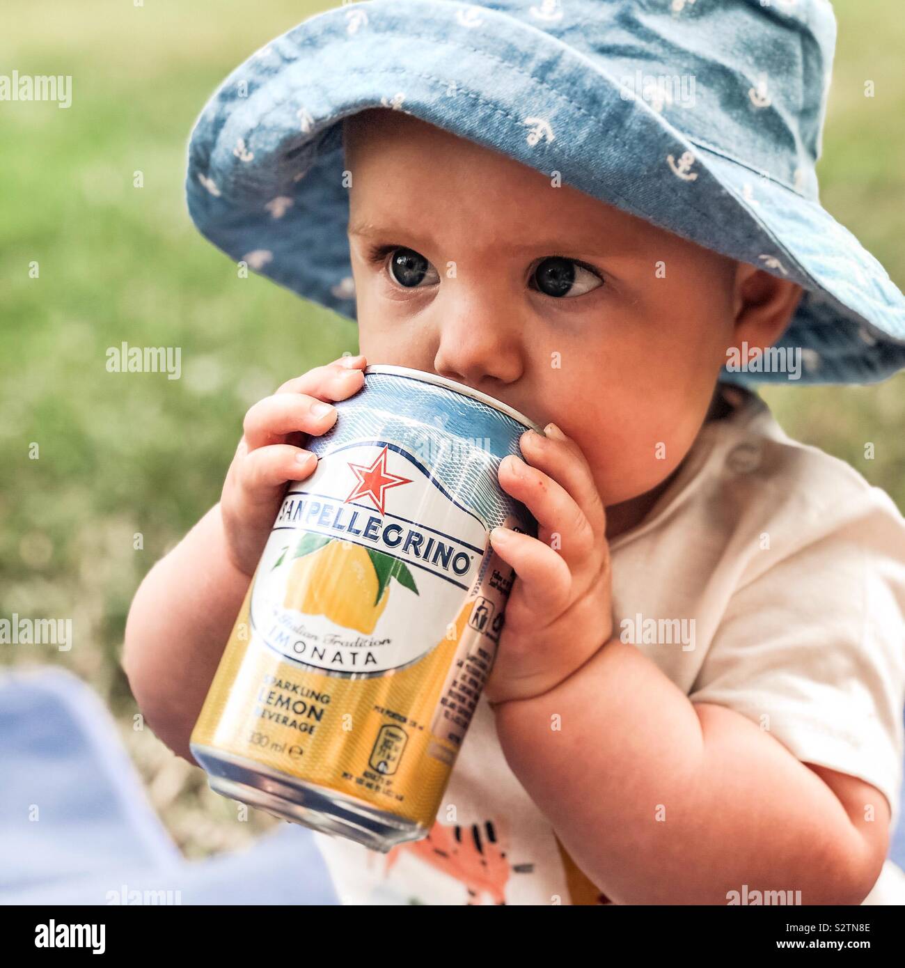 Baby boy, in the summer, drinking sanpellegrino lemonade can wearing a blue hat Stock Photo