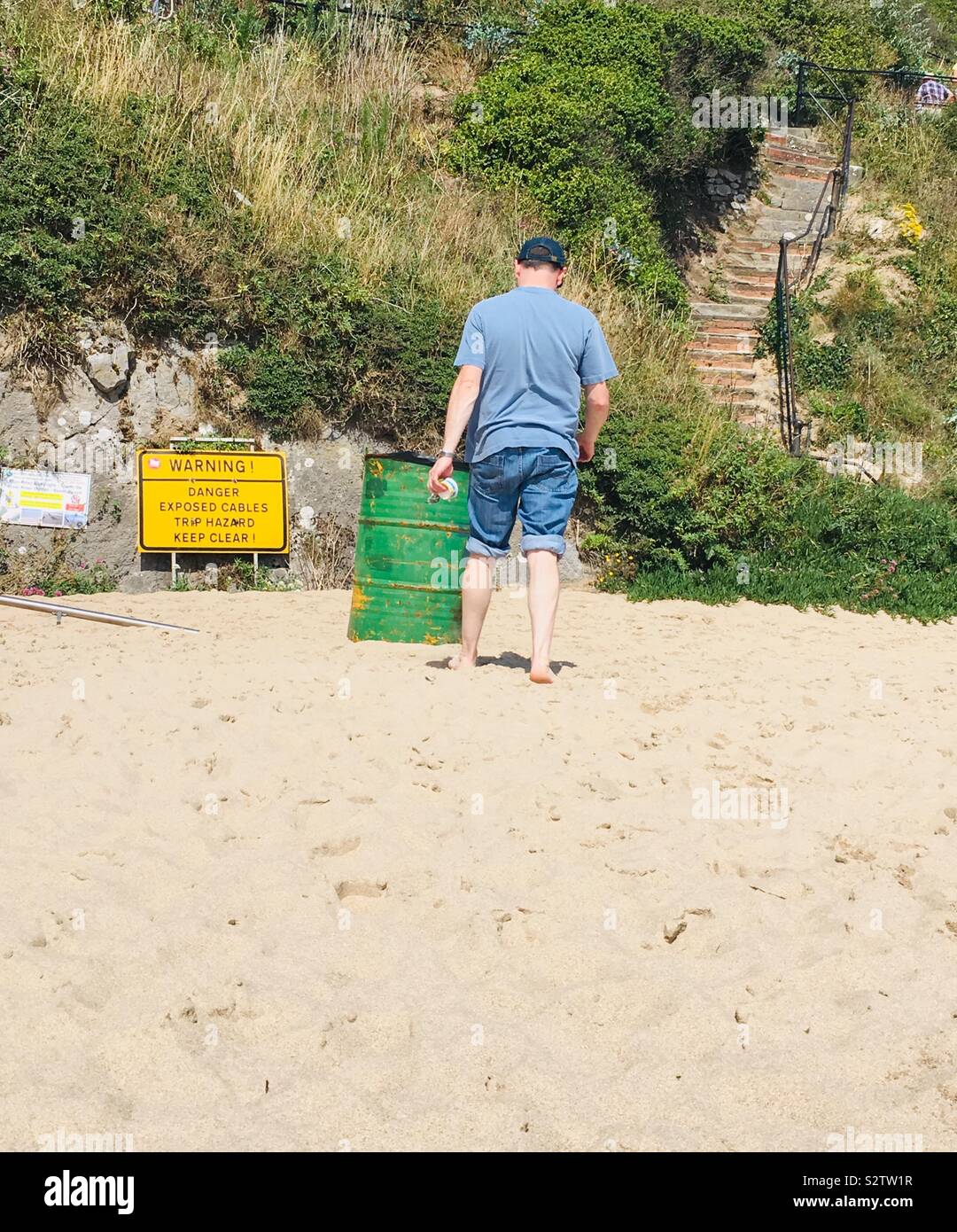Man wearing rolled up jeans, walking towards a bin and safety sign on a sandy beach with steps access Stock Photo