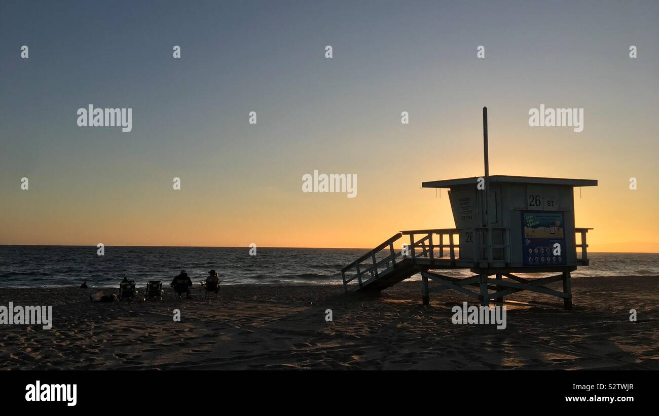 HERMOSA BEACH, CA, JUL 2019: silhouetted lifeguard hut and people sitting on chairs on shore next to Pacific Ocean at sunset - Smartphone Captured Stock Image