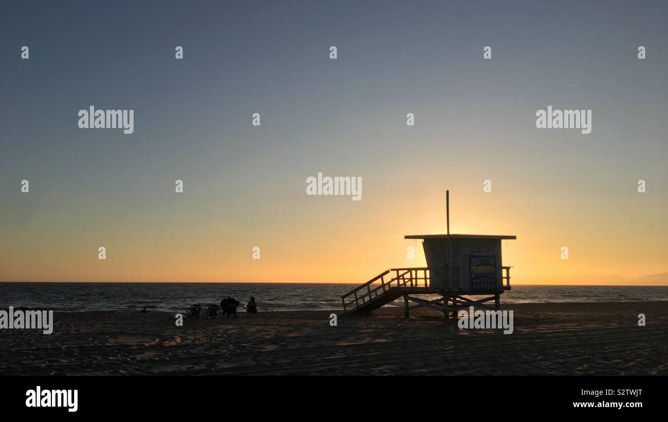 HERMOSA BEACH, CA, JUL 2019: silhouetted lifeguard hut and people sitting on chairs next to Pacific Ocean at sunset - Smartphone Captured Stock Image