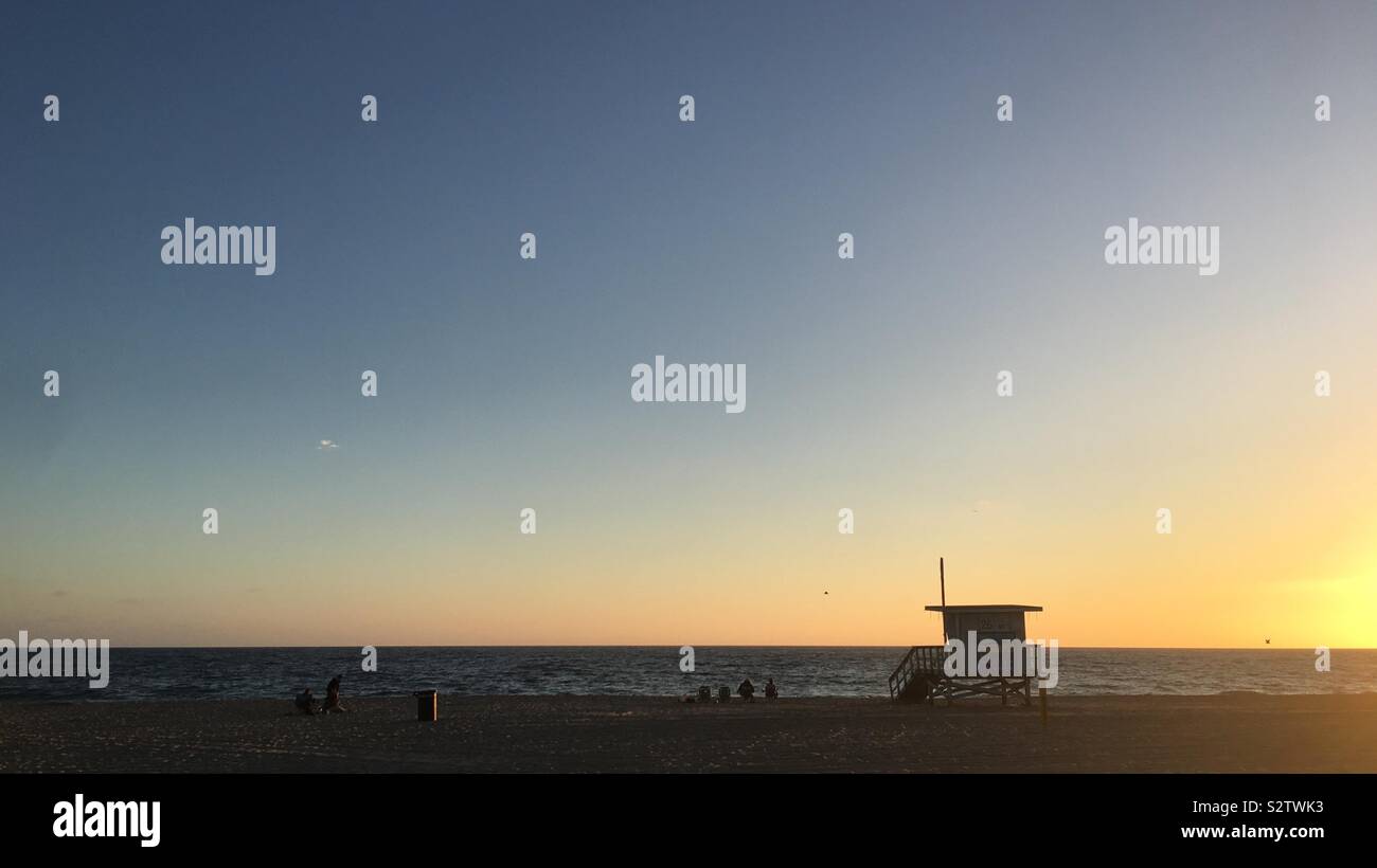 HERMOSA BEACH, CA, JUL 2019: wide view of people sitting next to Pacific Ocean, with sunset silhouetting them and nearby lifeguard hut - Smartphone Captured Stock Image