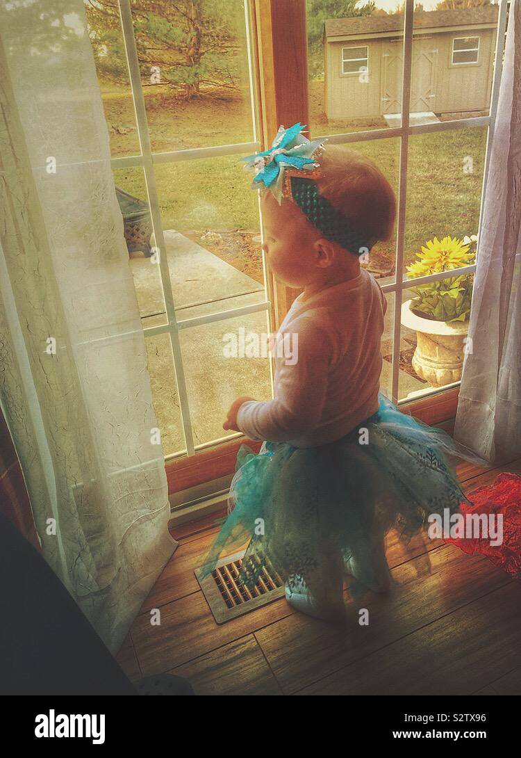 A baby girl, wearing a tutu skirt and bow headband, is looking out a sliding glass door. Stock Photo