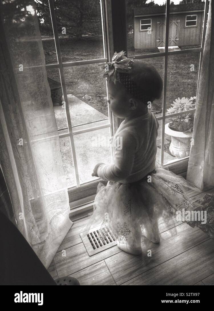 A baby girl, wearing a tutu skirt and bow headband, is looking out a sliding glass door.   A monochrome filter is used. Stock Photo
