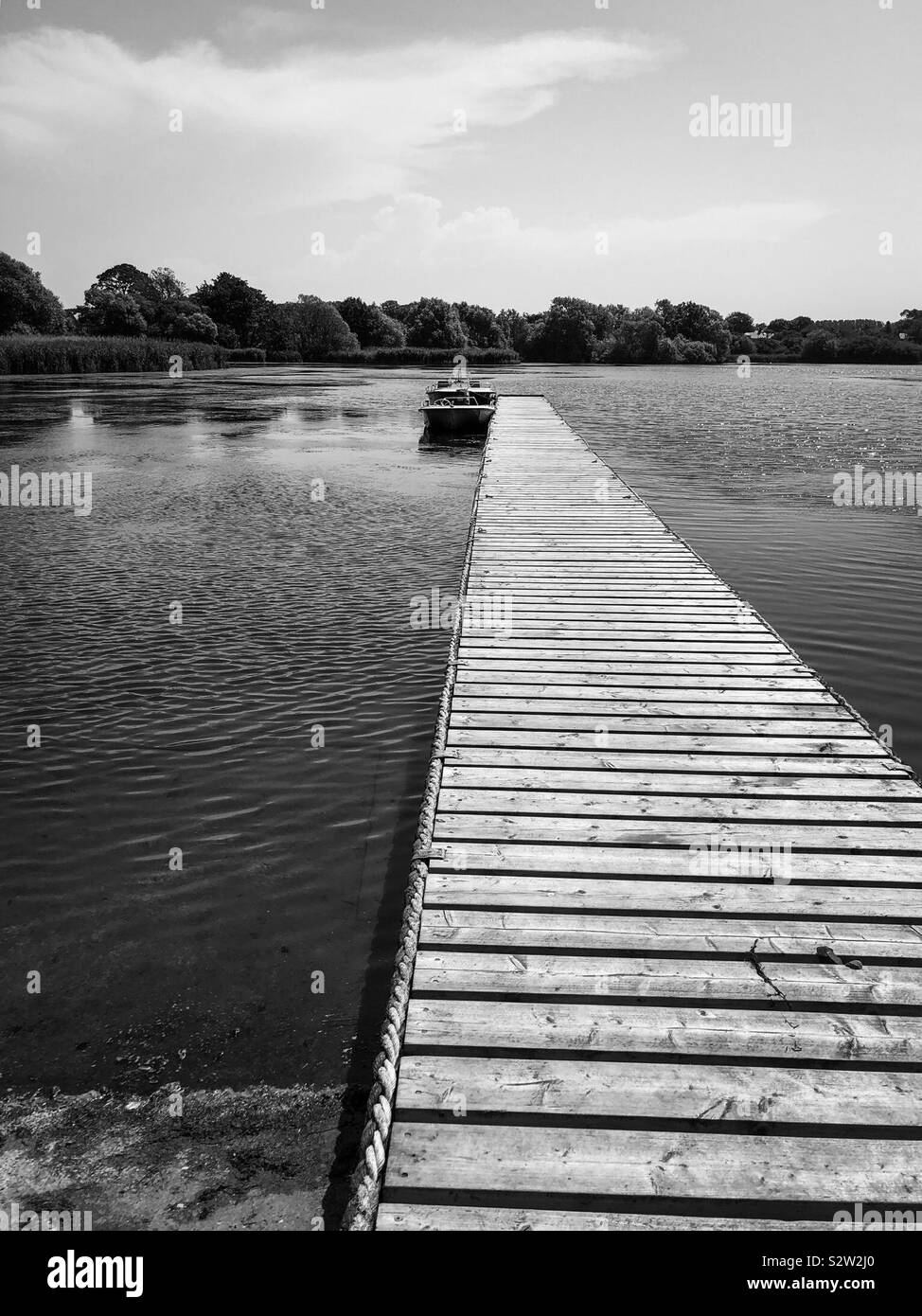 A black and white shot of a boat on a lake, moored beside a low, wooden jetty. - Smartphone Captured Stock Image