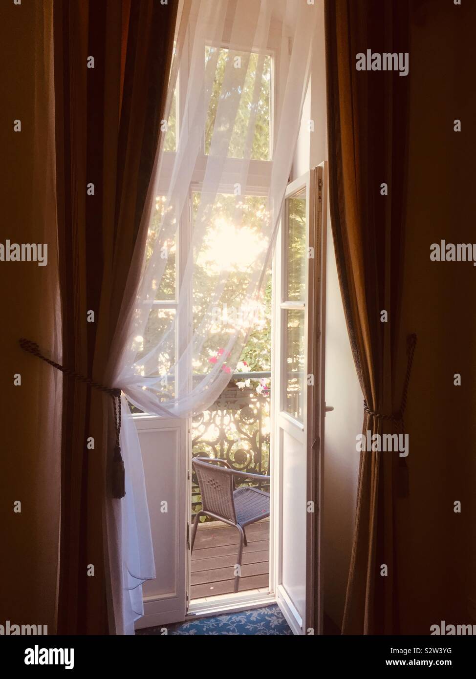 View through a balcony window with curtains of a chair and dappled sunlight. Stock Photo