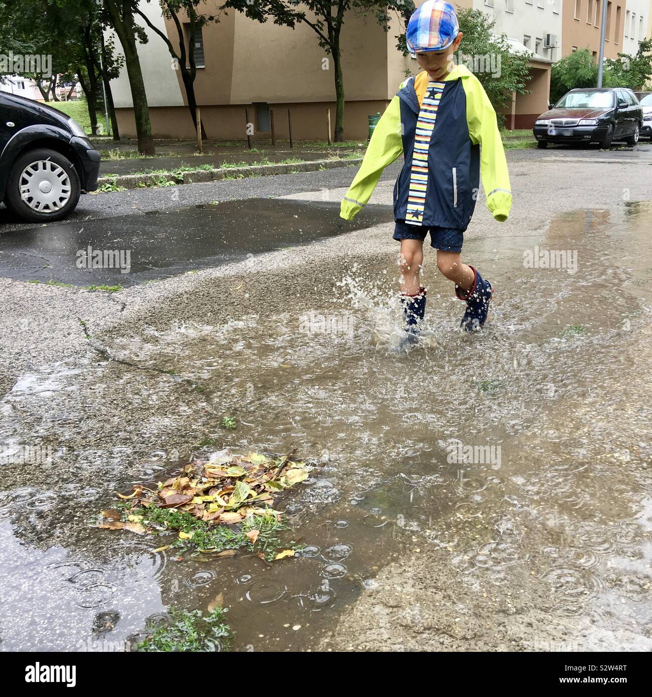 Child in raincoat wading in puddle after rain with drain grate blocked by leaves - Smartphone Captured Stock Image