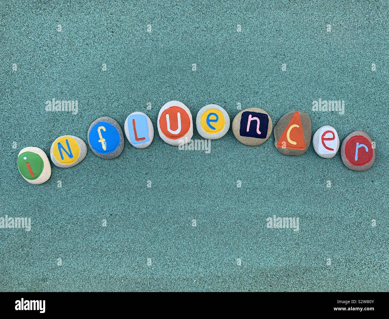 Influencer word composed with multi colored stone letters over green sand - Smartphone Captured Stock Image