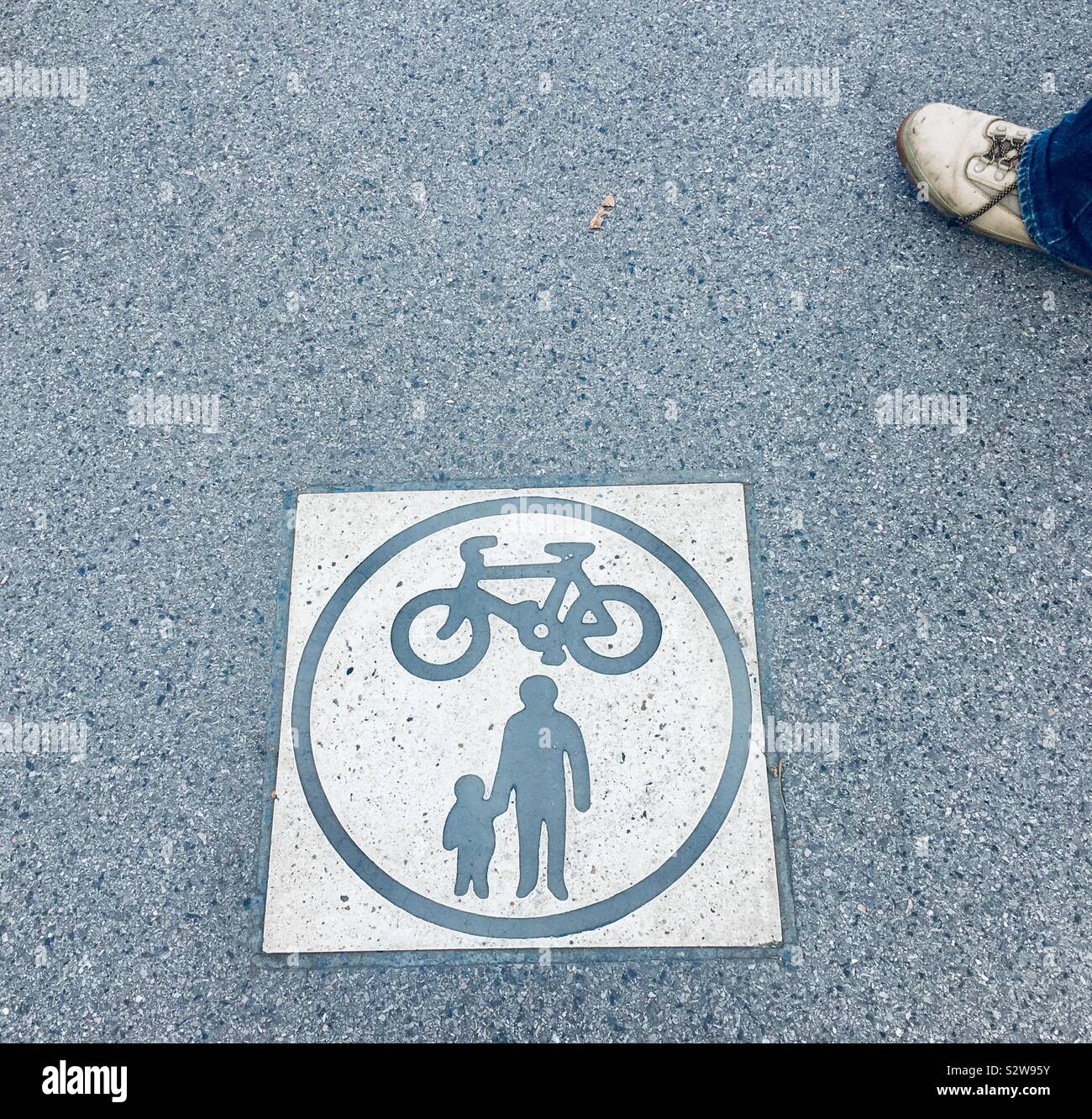 Aerial view of pair of a male's foot near traffic safety marking on tarred road - British pedestrian and cycling lane symbol - picture of a bicycle and a man with a child holding hands inside a circle Stock Photo