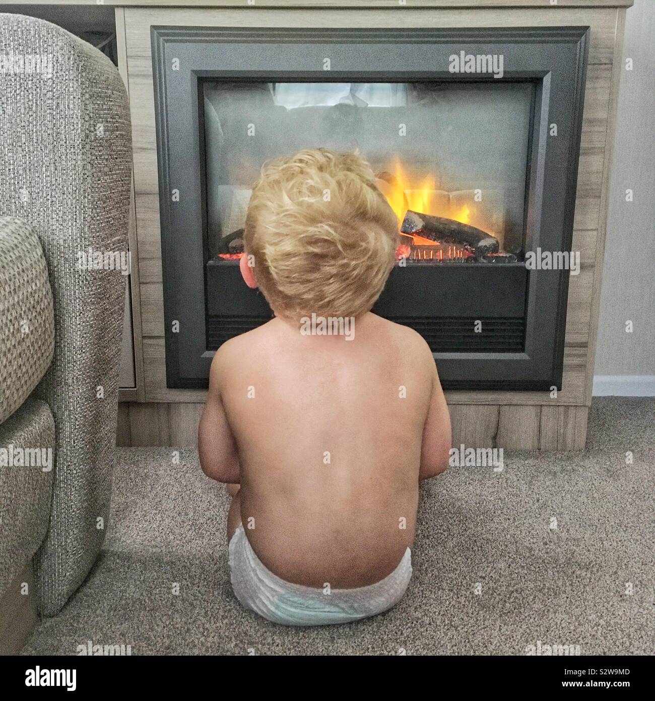 Two year old boy sitting near a fireplace, enjoying warmth in a cozy living room on a cold day - Smartphone Captured Stock Image