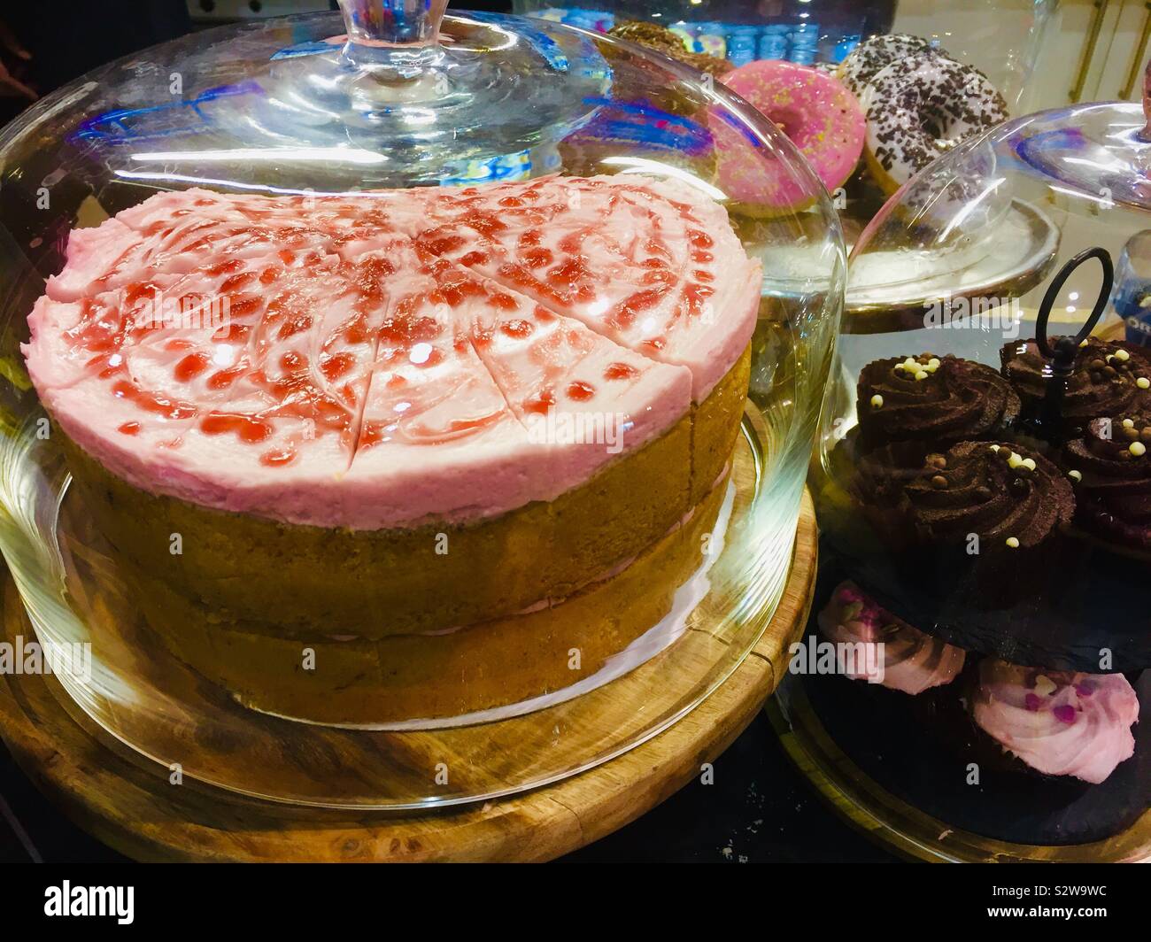 Cake display of a pink sponge cake under a glass dome, chocolate muffins and donuts filling the screen - Smartphone Captured Stock Image