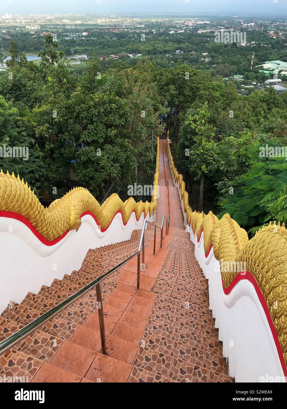 Steps with the Golden Naka statue on each side of the stairs leading down through a forest at Wat Phra That Doi Kham in Chiang Mai. Stock Photo