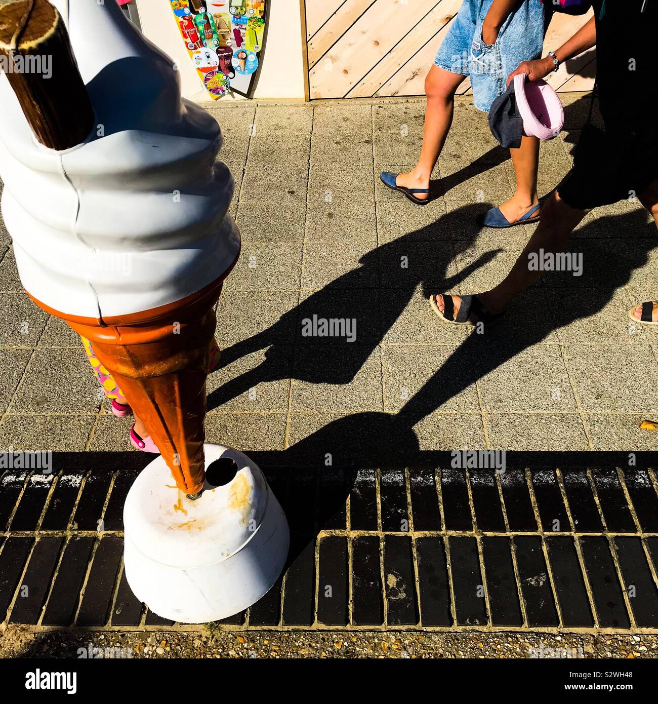 A beach seafront promenade scene showing feet legs and shadows of people walking past an ice cream kiosk large cone advertising model Stock Photo