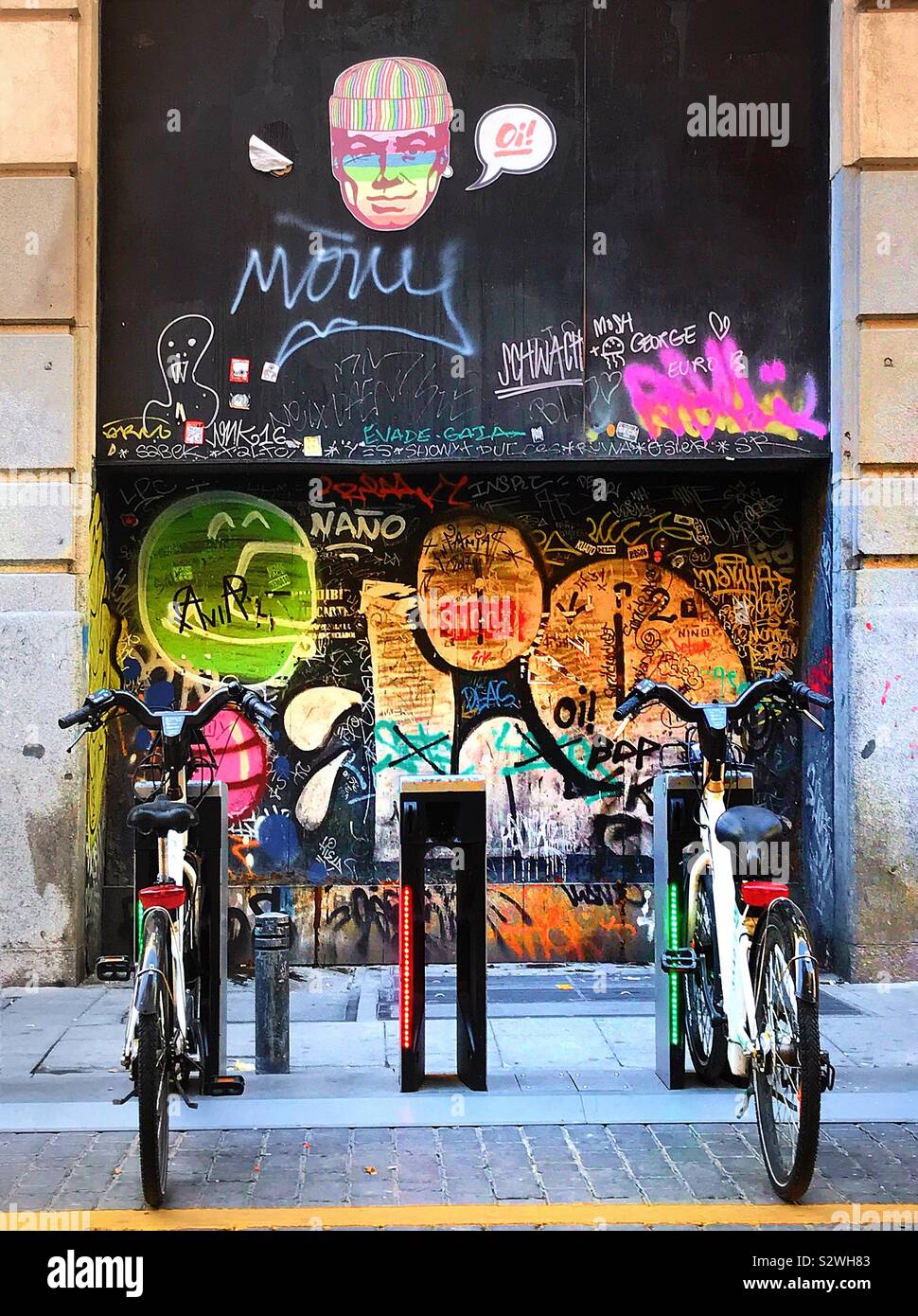 Electric bicycles in their docking stations in Madrid’s trendy district of Malasaña. In the background the wall is covered with an array of graffiti and stickers. Stock Photo