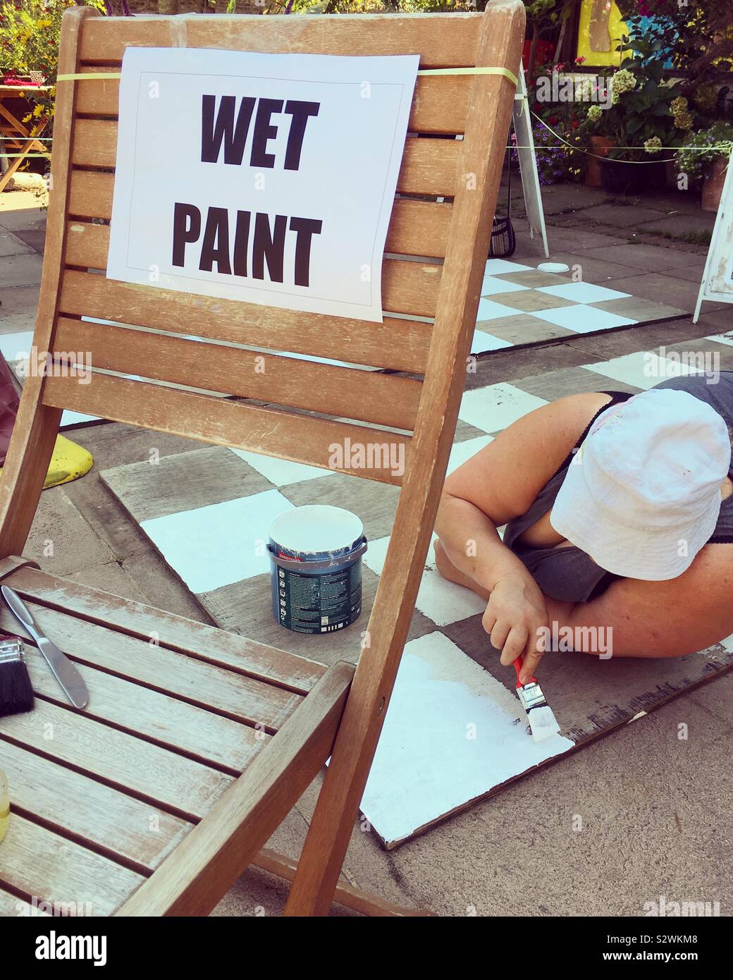 Wet paint sign alongside a woman who is painting an oversize oversized black and white chessboard with white paint on the white squares, in the street. Twickenham, UK. - Smartphone Captured Stock Image