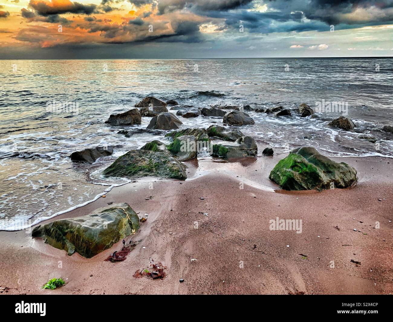 Moody Ocean. The incoming tide laps around large boulders on the shore. A stormy sky reflects the golden hues of the sunset. - Smartphone Captured Stock Image